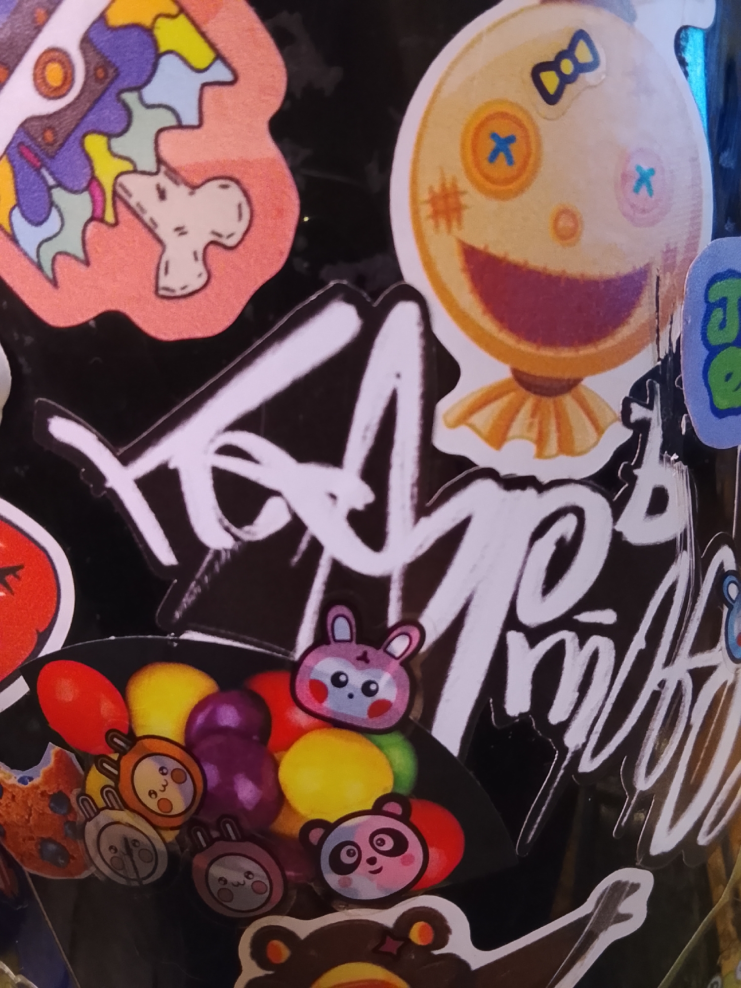The image depicts a collection of stickers adhered to a black surface. The stickers include a stylized brain, a ragdoll-like face with buttons for eyes and a bow, a plate of fruits and animal-faced candies, and a large signature written in white. The location is Высокиничи, Russia.