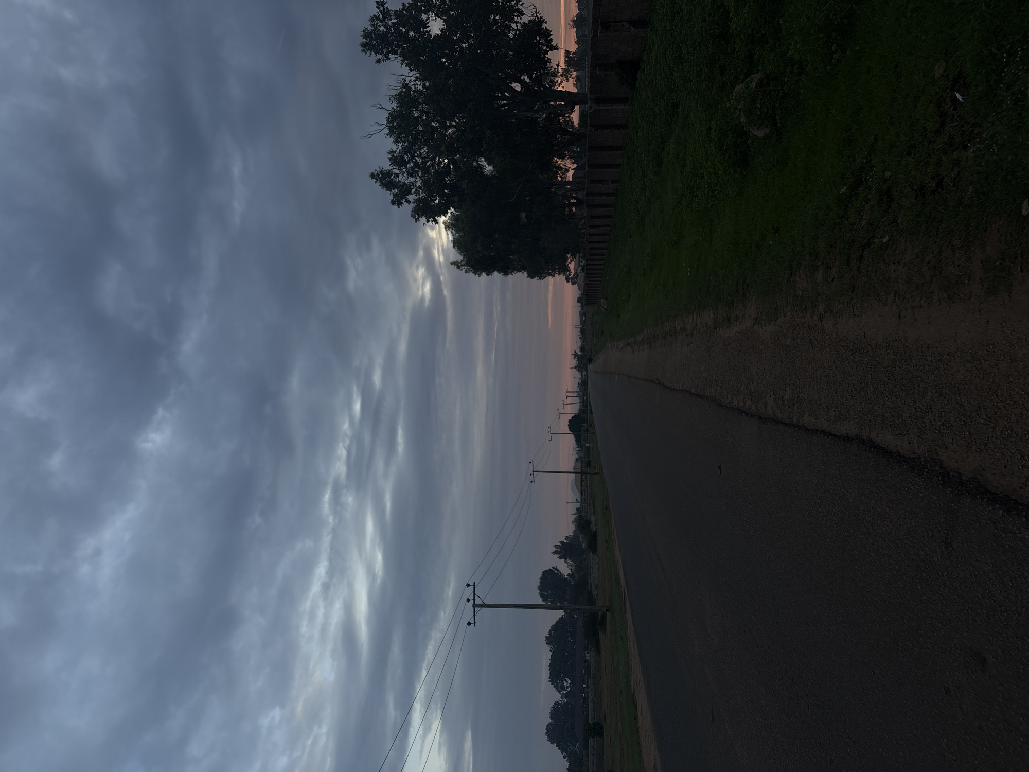 The image displays a deserted road scene in Bukuru, Nigeria, during either sunrise or sunset, characterized by an overcast sky. Dense, grey and blue-grey stratocumulus clouds dominate the sky, with a horizontal band of soft orange and pink light visible low on the horizon, particularly prominent to the right.
A dark asphalt road extends from the foreground into the distance, appearing to be a two-lane thoroughfare without markings, slightly curving leftward. A narrow dirt shoulder borders the immediate right edge of the asphalt.
On the left side of the road, a series of wooden utility poles with multiple power lines recede into the background. Beyond these poles, clusters of dark green trees and dimly visible, low-rise structures are present in the mid-ground.
On the right side of the road, a strip of green grass and low-lying vegetation runs parallel to a dirt path that serves as a wider shoulder. Further right, a weathered, low-height concrete or stone perimeter wall stretches into the distance. Behind this wall, a line of mature, dark-silhouetted trees stands against the lighter horizon, with the closest tree exhibiting significant foliage and some bare branches.
No individuals, vehicles, or animals are visible, rendering the scene tranquil and devoid of immediate activity. The environment suggests a semi-rural or outer-urban area of Bukuru.