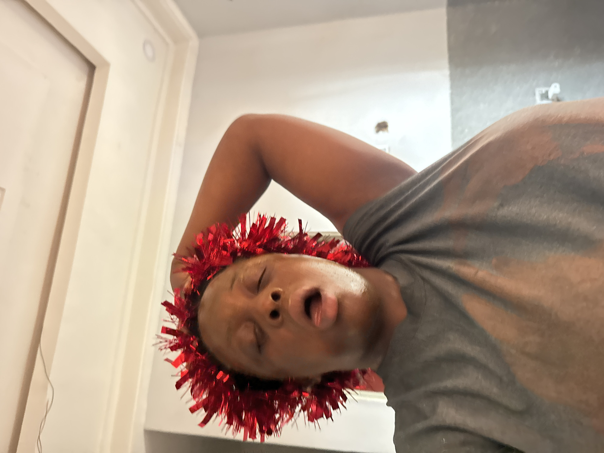 A person with dark skin is wearing a red tinsel garland around their head. Their head is tilted back and their mouth is open. They are wearing a gray t-shirt. The background includes a white door and a gray wall. There is no evidence to indicate the location is Sabon Tasha, Nigeria.