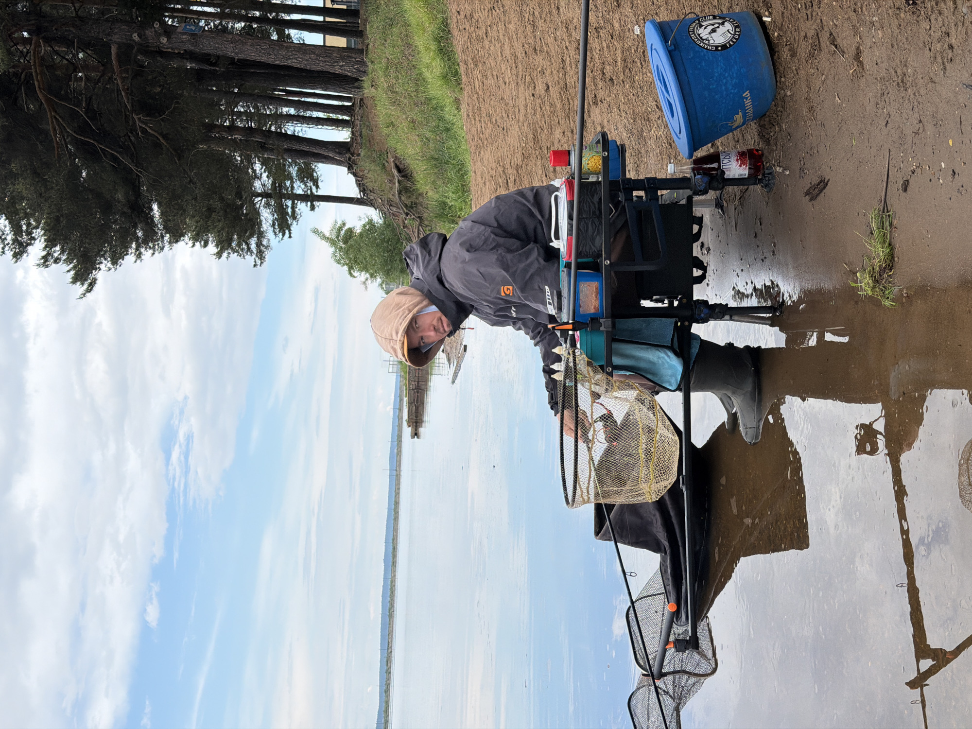 A fisherman in Chaikovsky, Russia, prepares his gear on the bank of a body of water. The man is wearing a hooded jacket and boots. He is seated on a fishing platform with equipment. A blue bucket and a bottle are visible near him on the ground. Fishing rods and nets are positioned around the platform. Trees line the bank in the background, with a view of the water and sky in the distance.