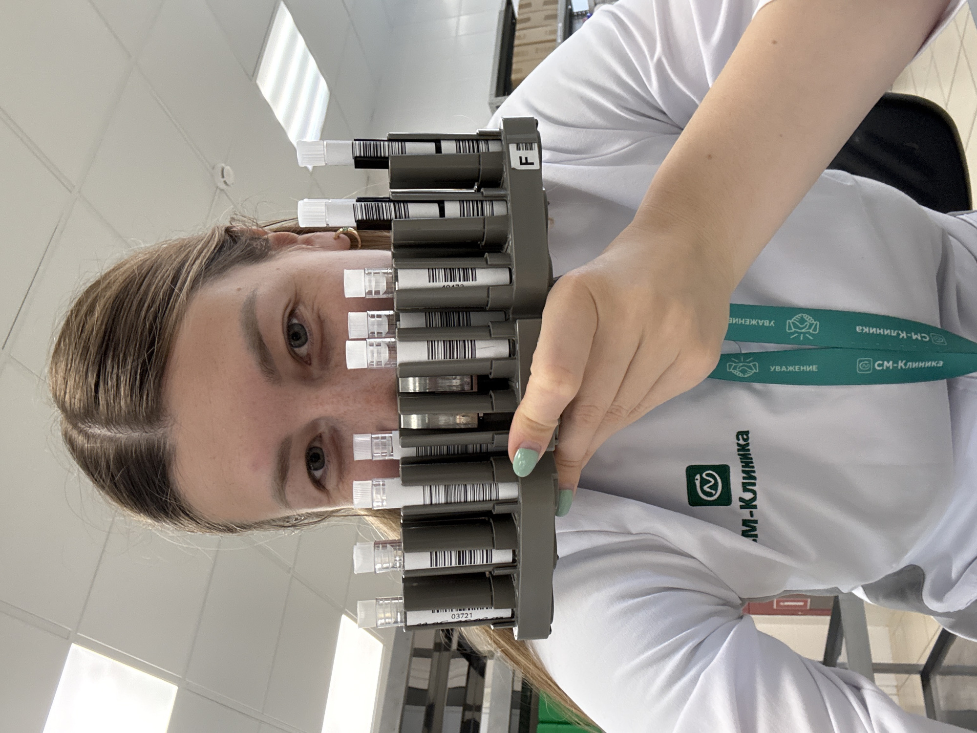 The image features a woman holding a rack of test tubes in front of her face. She has fair skin, light brown hair, and blue eyes. She is wearing a white shirt with the logo "CM-Клиника" on it, and a green lanyard with the same logo is visible around her neck. The test tubes in the rack have white caps and barcode labels. The background is a bright, sterile-looking room. Based on the logo on the shirt and lanyard, the location is likely in Moscow, Russia, as "CM-Клиника" is a chain of medical clinics in that city.