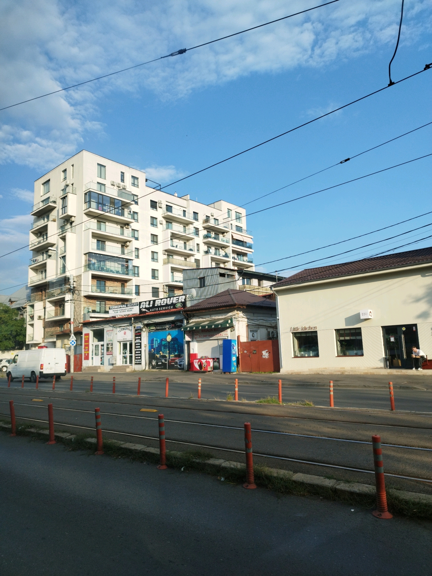 The image depicts a street scene in Bucharest, Romania, under a bright blue sky with scattered clouds. The foreground features an asphalt road with tram tracks and a row of orange bollards with grey reflective bands. A white van is parked on the side of the road. To the right of the van, several businesses are visible. One building is a modern, multi-story apartment complex with balconies. Next to it are single-story commercial buildings. One of these houses a business with a sign that reads "ALI ROUVER AUTO SERVICE." Further down the street, a building with a sign "Little Kitchen" is visible. A person is seated outside this establishment. The overall impression is a typical urban environment with commercial and residential buildings, infrastructure for public transportation, and everyday street life.