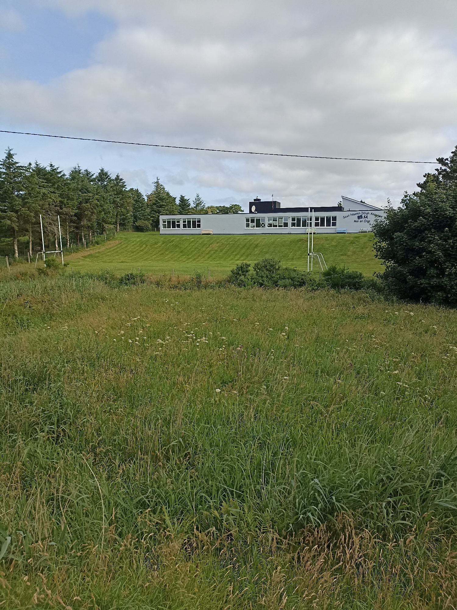 A school building, "Scoil Naomh Duigh An Ghlase," is situated on a grassy hill in Annagry, Ireland. The building is modern, with a grey facade and large windows. In front of the building, a neatly mown field slopes down, featuring a set of goalposts. Beyond the mown field, a dense line of trees forms the backdrop. In the foreground, a wild, overgrown meadow is visible, filled with tall grass and scattered white flowers. The sky above is partly cloudy. The image captures a rural school environment on a slightly overcast day.