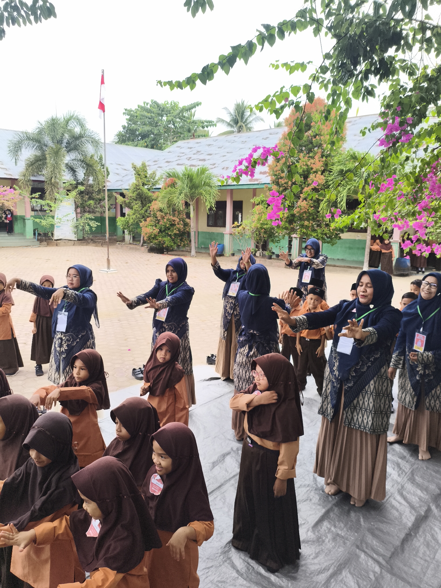 In Baratpayaitek, Indonesia, a group of school children and adults are gathered outdoors on a paved area, possibly a schoolyard. The children, predominantly girls wearing brown hijabs and orange uniforms, are positioned in front, some seated, others standing. Adults, dressed in dark blue tops with intricate patterns and tan skirts, appear to be instructors or guardians, leading the children in what seems to be a synchronized activity or dance. Their hands are raised and positioned in a similar fashion, suggesting a coordinated movement. Behind the main group, more children in similar attire are visible, some looking towards the activity, while others are partially obscured. The background features a low-rise building with a light-colored roof, palm trees, and vibrant pink bougainvillea flowers hanging from a structure, indicating a tropical or subtropical environment. A flagpole with the Indonesian flag is also present in the distance. The overall scene depicts an organized educational or recreational event taking place in an outdoor school setting.
