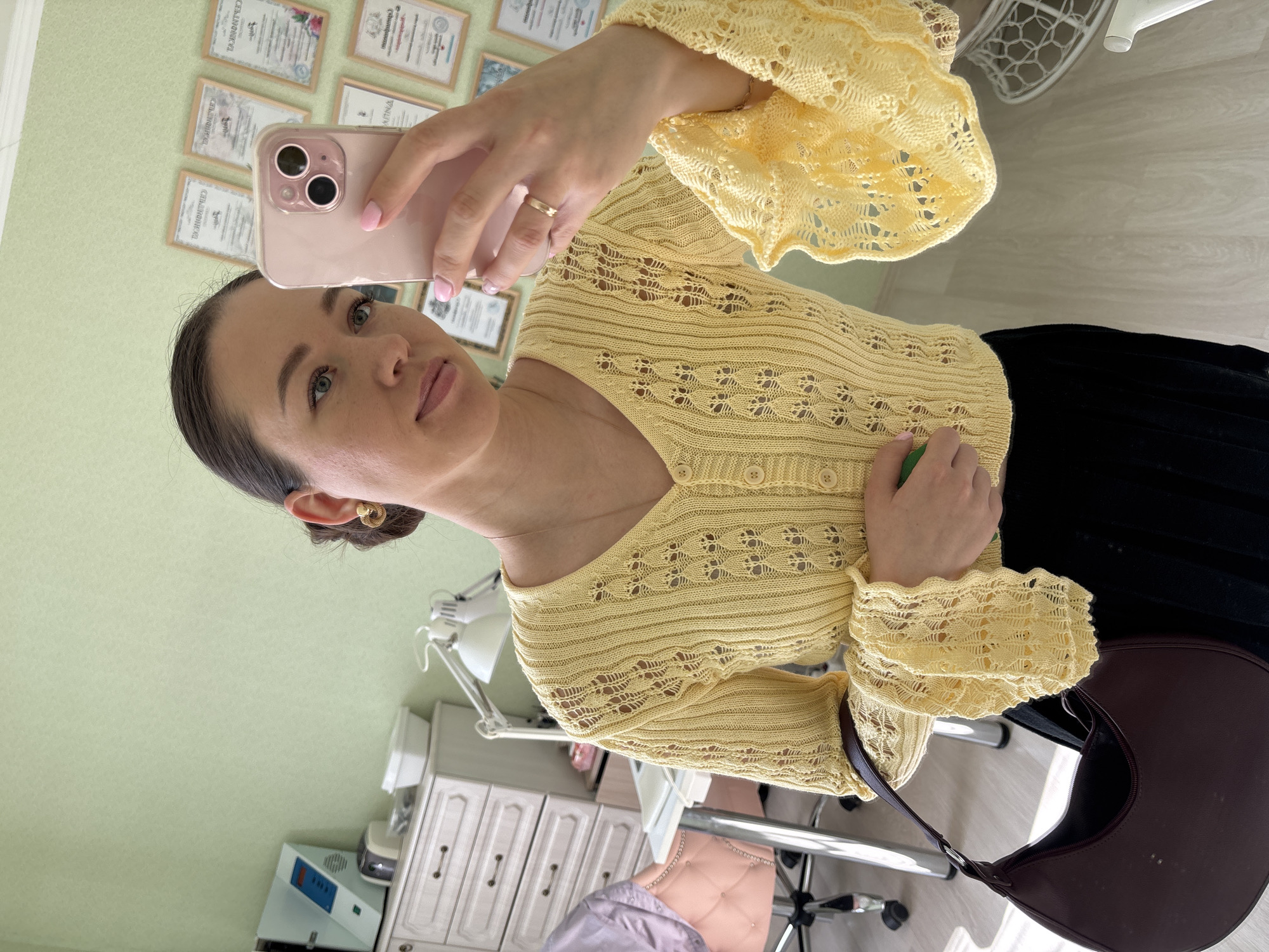 A woman takes a selfie in a room in Ферма, Russia. She is wearing a yellow knitted sweater and a dark-colored skirt. She is holding a pink smartphone, and there is a gold ring on her finger. A brown purse is visible. The background includes a wall with framed certificates, a lamp, a manicure device, and cabinets.
