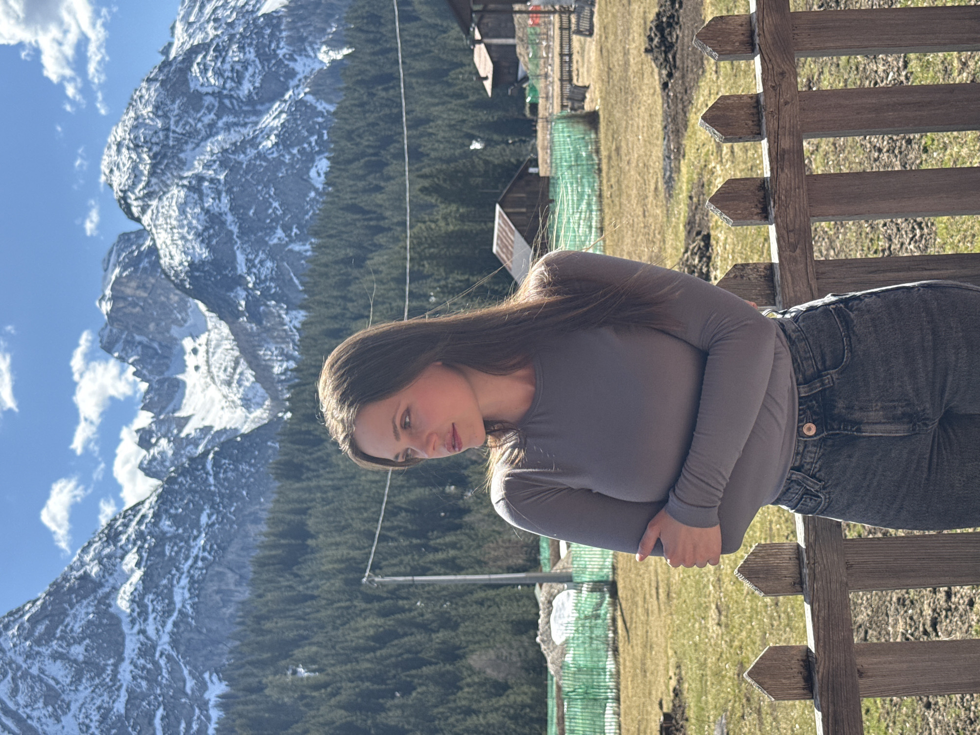 A woman is sitting on a wooden fence with snow-capped mountains in the background in Sappada, Italy. The woman has long brown hair and is wearing a gray long-sleeved shirt and jeans. She is looking to the left, away from the camera. The background shows a lush green forest and a bright blue sky with some clouds. There are buildings in the distance. The woman is posed near a wooden fence in the foreground.
