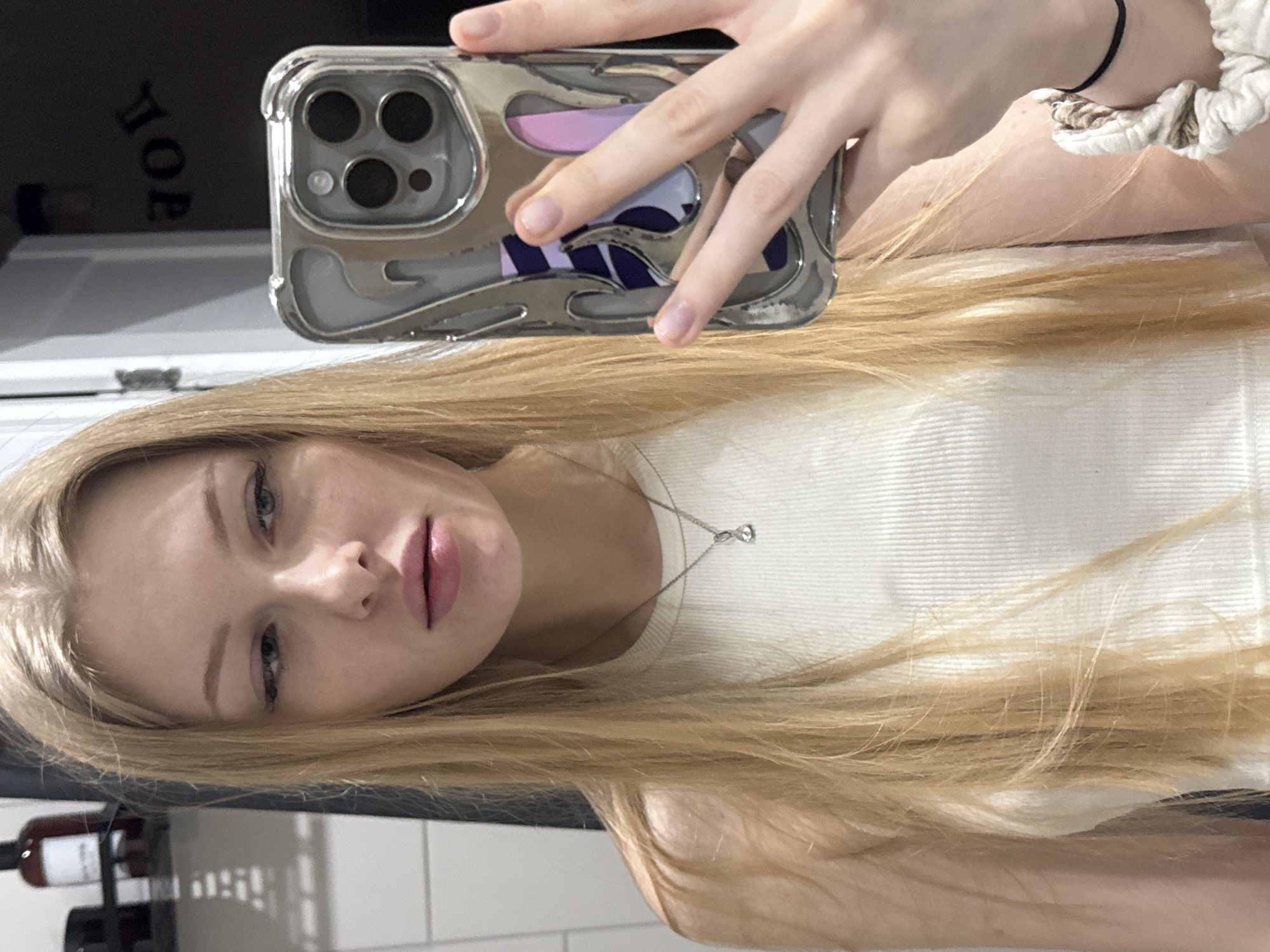 A blonde woman is taking a selfie. She is holding a silver iPhone with a clear case. She is wearing a white tank top and a silver necklace. She has a scrunchie on her wrist. In the background, there is a bathroom with white tiles and a bottle of wine on the counter. There is no indication of the city of Perm, Russia, or any other specific location.