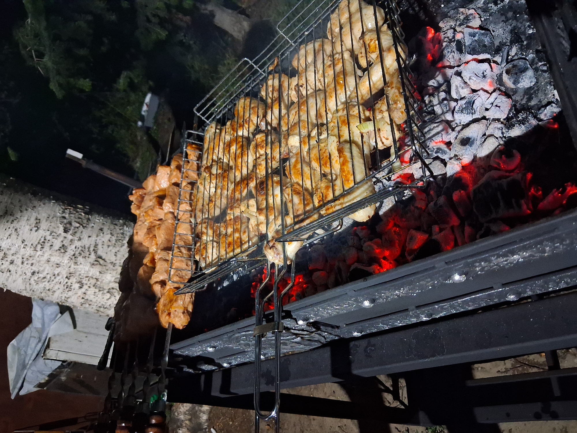 An outdoor night scene in Semey, Kazakhstan, features a barbecue with a grill full of chicken pieces and raw meat skewers. The grill is placed over glowing red coals, indicating active cooking. In the background, a large tree with white bark is visible, along with dark foliage and some scattered debris. The overall ambiance suggests a casual gathering or a backyard cookout.
