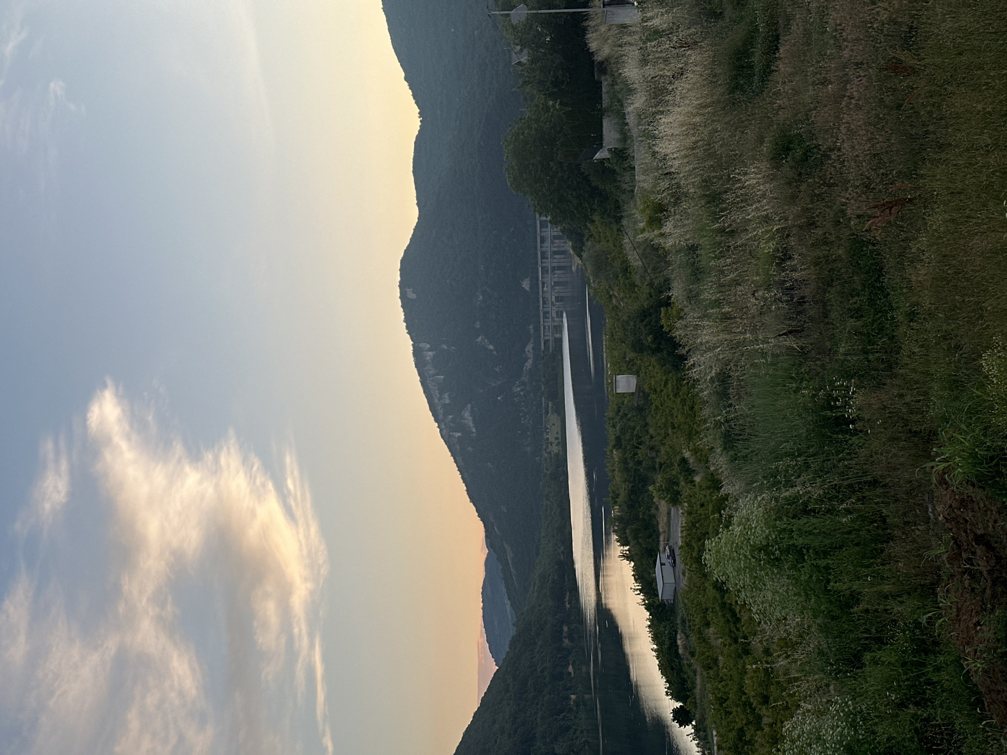 This is a wide-angle, landscape photograph captured at an outdoor location, likely during the golden hour or twilight. The scene depicts a vast body of water, possibly a lake or reservoir, surrounded by lush, tree-covered hills. In the distance, a large concrete structure, possibly a dam or bridge, spans across the water. The sky above is a gradient of soft blues and oranges, with scattered clouds catching the warm light. The foreground is dominated by tall, dry grasses and other vegetation, creating a textured, natural foreground. A small white building with a dark-colored vehicle parked in front of it is visible on the shoreline to the left of the frame, nestled amongst the trees. The overall atmosphere is serene and tranquil, with a sense of natural beauty and vastness.