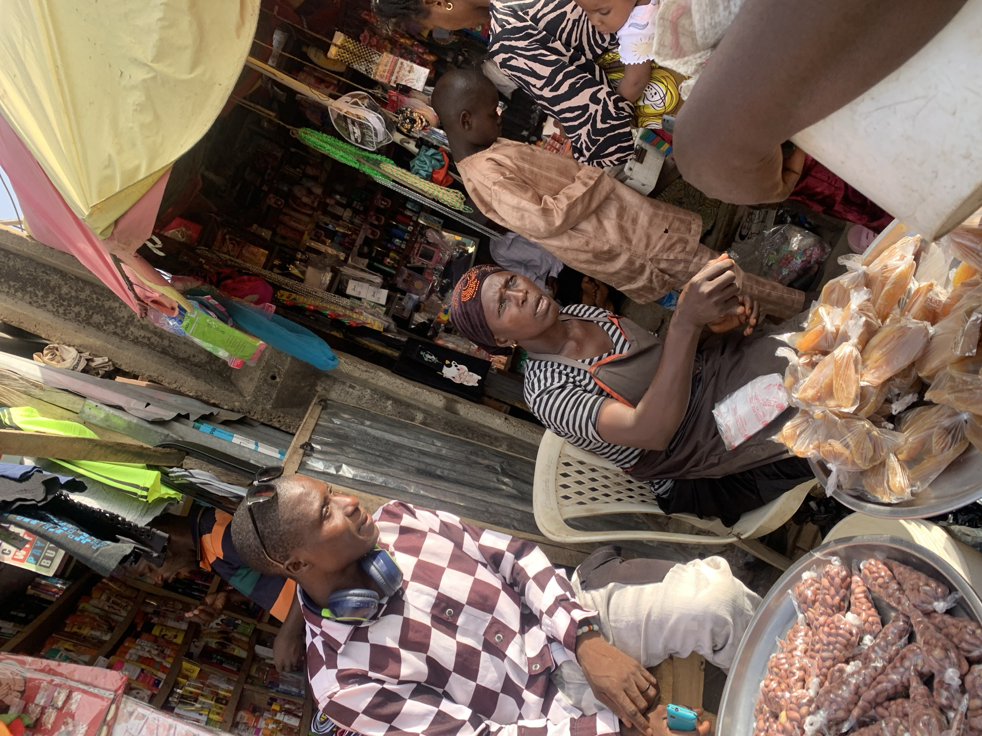 This image captures a vibrant, bustling outdoor market scene in Jos, Nigeria, under bright daylight with strong shadows indicating sunny weather. The perspective is slightly overhead, looking down into various stalls and activities.
In the foreground, a man sits relaxed, wearing a maroon and white checkered long-sleeve shirt and light trousers. He has blue headphones around his neck and sunglasses pushed onto his head, looking up and to the right. To his right, two large metal trays are filled with individually packaged snacks: one tray holds what appears to be fried groundnuts in clear plastic bags, and the other contains similar-looking items, possibly another local delicacy.
In the mid-ground, a woman with a serious or weary expression sits on a white plastic chair, wearing a striped shirt and a dark headwrap with orange details. She holds something in her hands, possibly a small bottle. Behind her and extending into the background, improvised market stalls are visible, constructed with corrugated metal roofs and wooden supports. These stalls are packed with a wide variety of goods on shelves, including packaged foods, toiletries, and other consumer products. Green, woven ropes or beads hang prominently in one section.
Further in the background, a young boy in a light brown traditional top looks to the right. Next to him, an adult in a black and white zebra-striped top holds a small child dressed in yellow patterned clothing. Overhead, a yellow and pink fabric awning provides shade.
The scene depicts the daily life of a busy market. The people are engaged in selling, waiting, or observing. Visible text is limited, but "VIVA WAFER" can be partially seen on a white plastic bag, and "C PAY BUY" on a sign in the background shelves. The informal setup and variety of goods are strong cues of a local market environment.