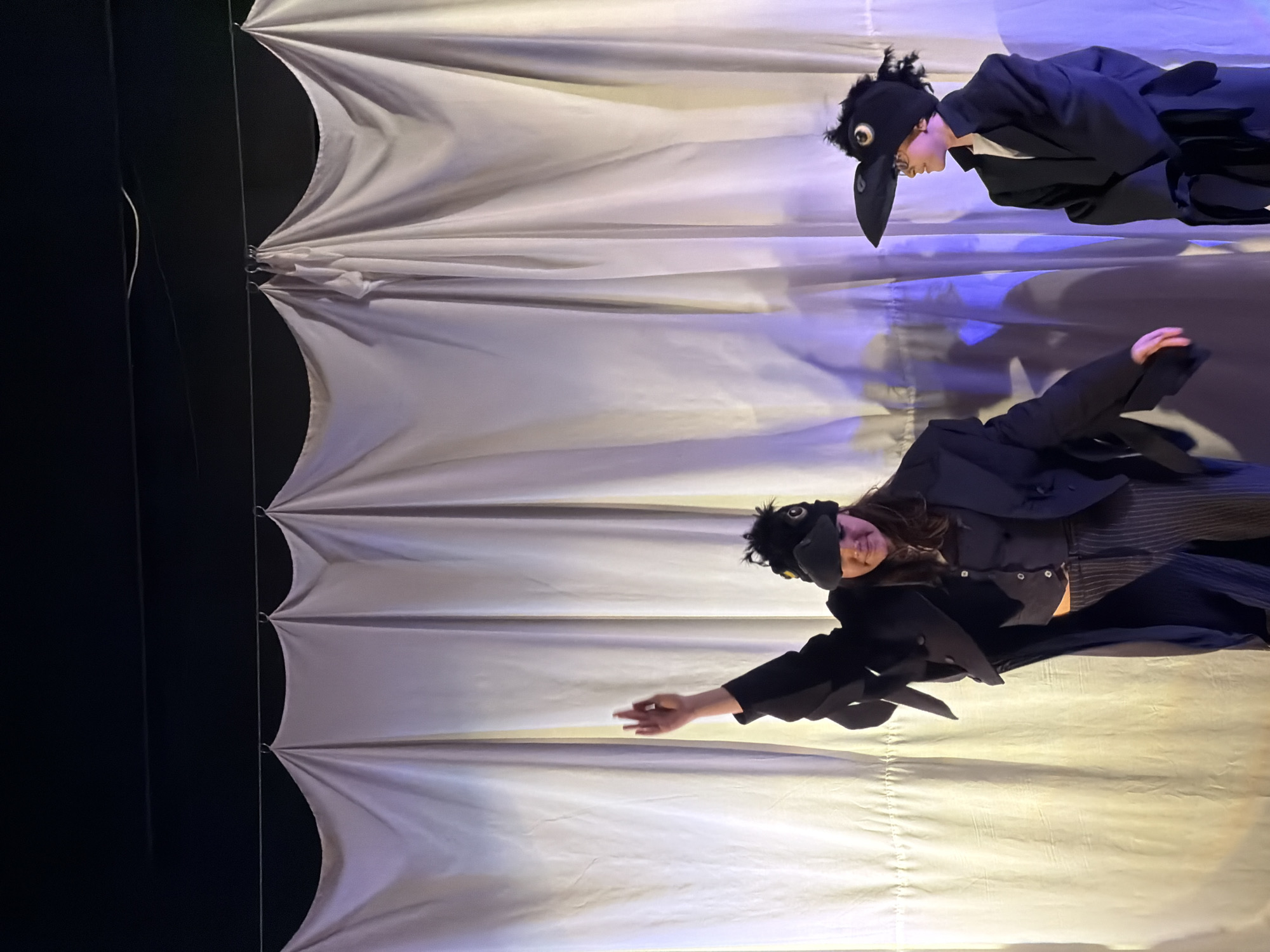 Two people wearing crow costumes are positioned in front of a white, vertically hanging curtain. The curtain is bunched in evenly spaced intervals. The background is dark. The image was taken in the city of Pleven, Bulgaria.