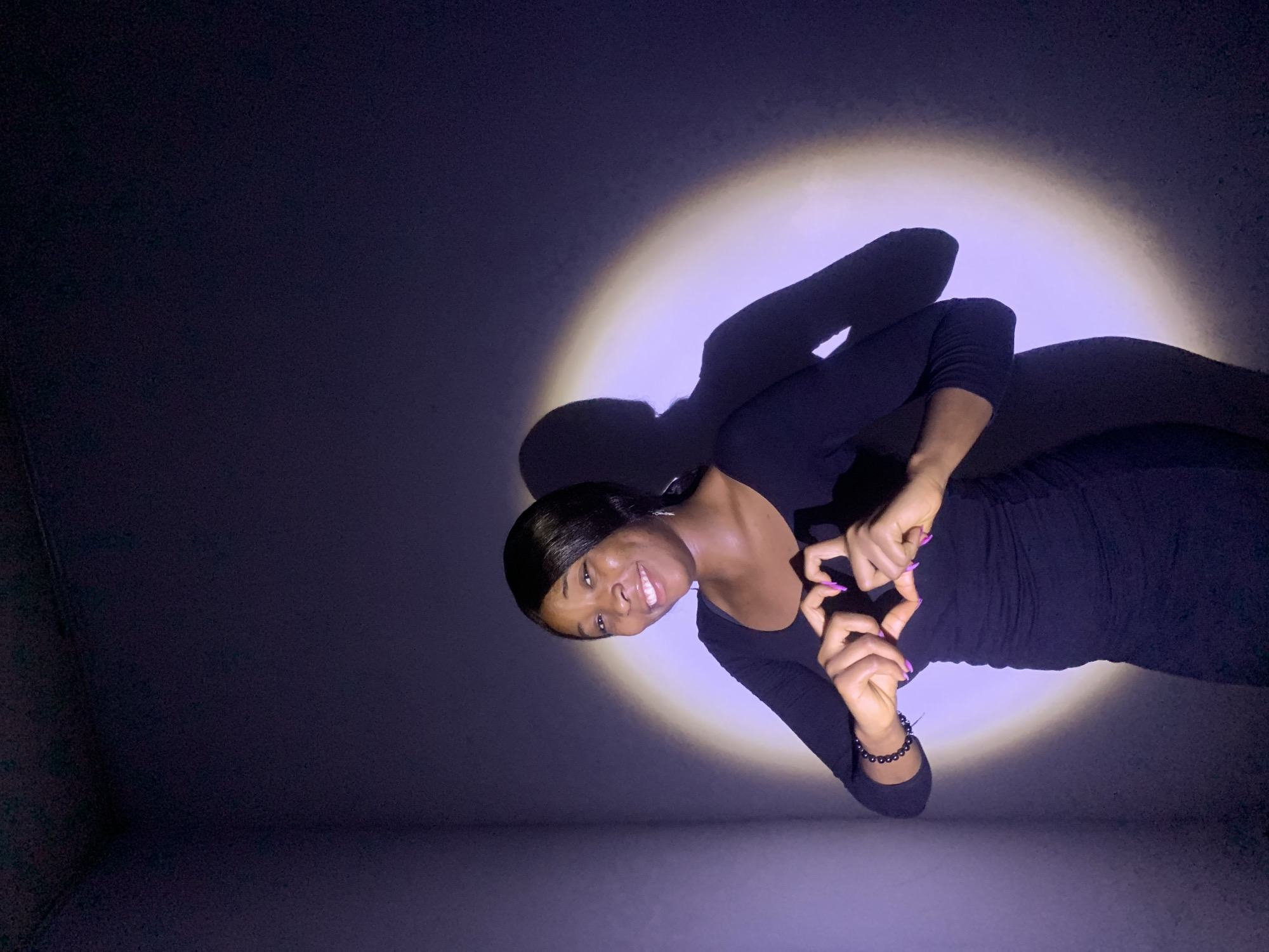 A medium shot of a Black woman, facing the camera and smiling, is the central focus. She is positioned on a stark gray floor against a dark gray backdrop, illuminated by a single, off-center light source creating a halo effect behind her. The woman is wearing a long-sleeved black dress and a bracelet on her left wrist. Her nails are painted pink. She holds her hands together to form a heart shape. The environment is minimal, consisting of a dark gray background and a lighter gray floor, suggesting an indoor space. There is no indication of Jos, Nigeria or any city location.