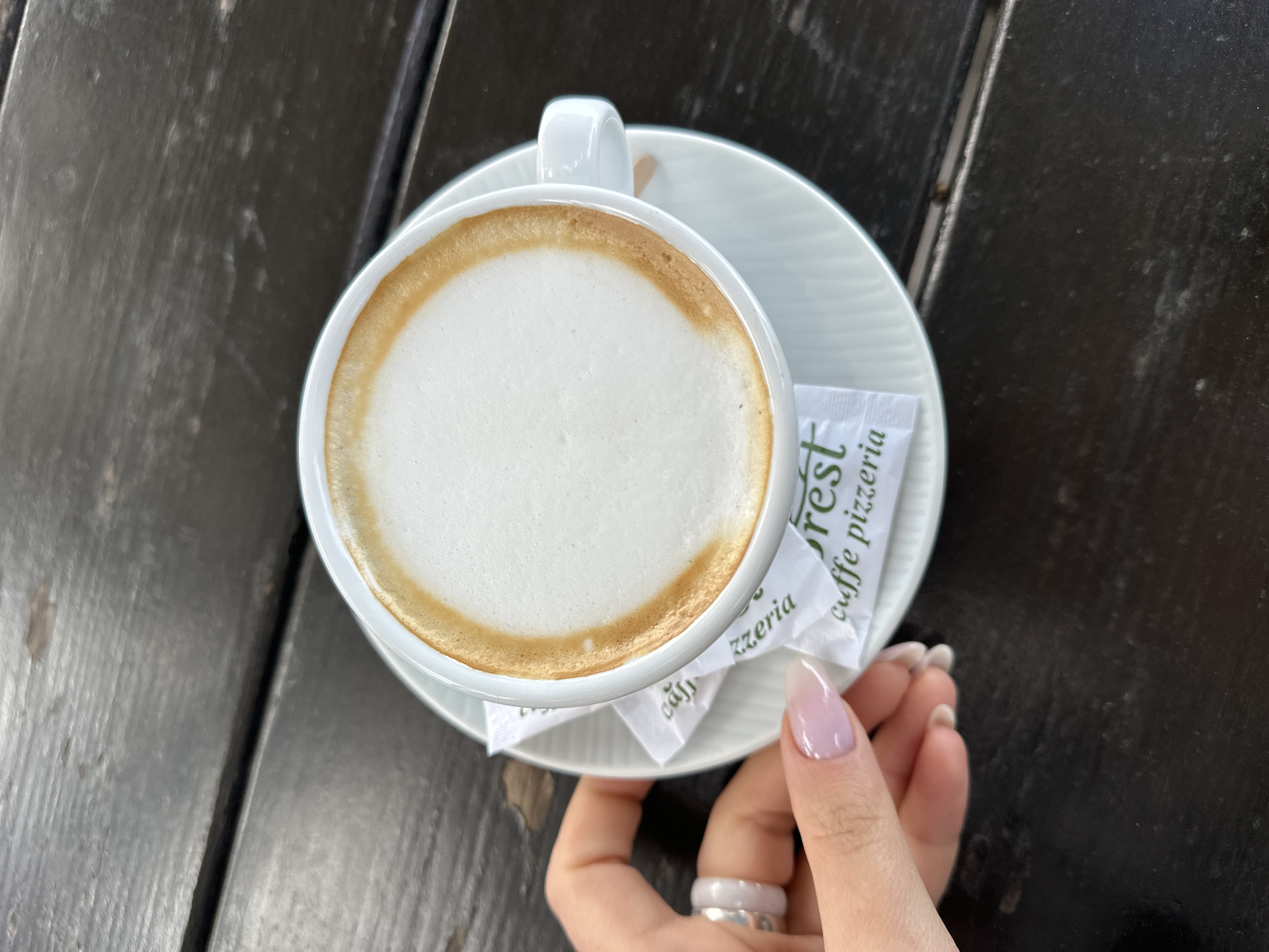 An overhead shot shows a white cup of cappuccino on a matching saucer, set on a dark wooden table. Two packets of sugar, labeled "Forest caffe pizzeria," rest on the saucer next to the cup. A hand, adorned with a silver ring and light pink manicure, holds the saucer. The setting is Šušanj, Montenegro.