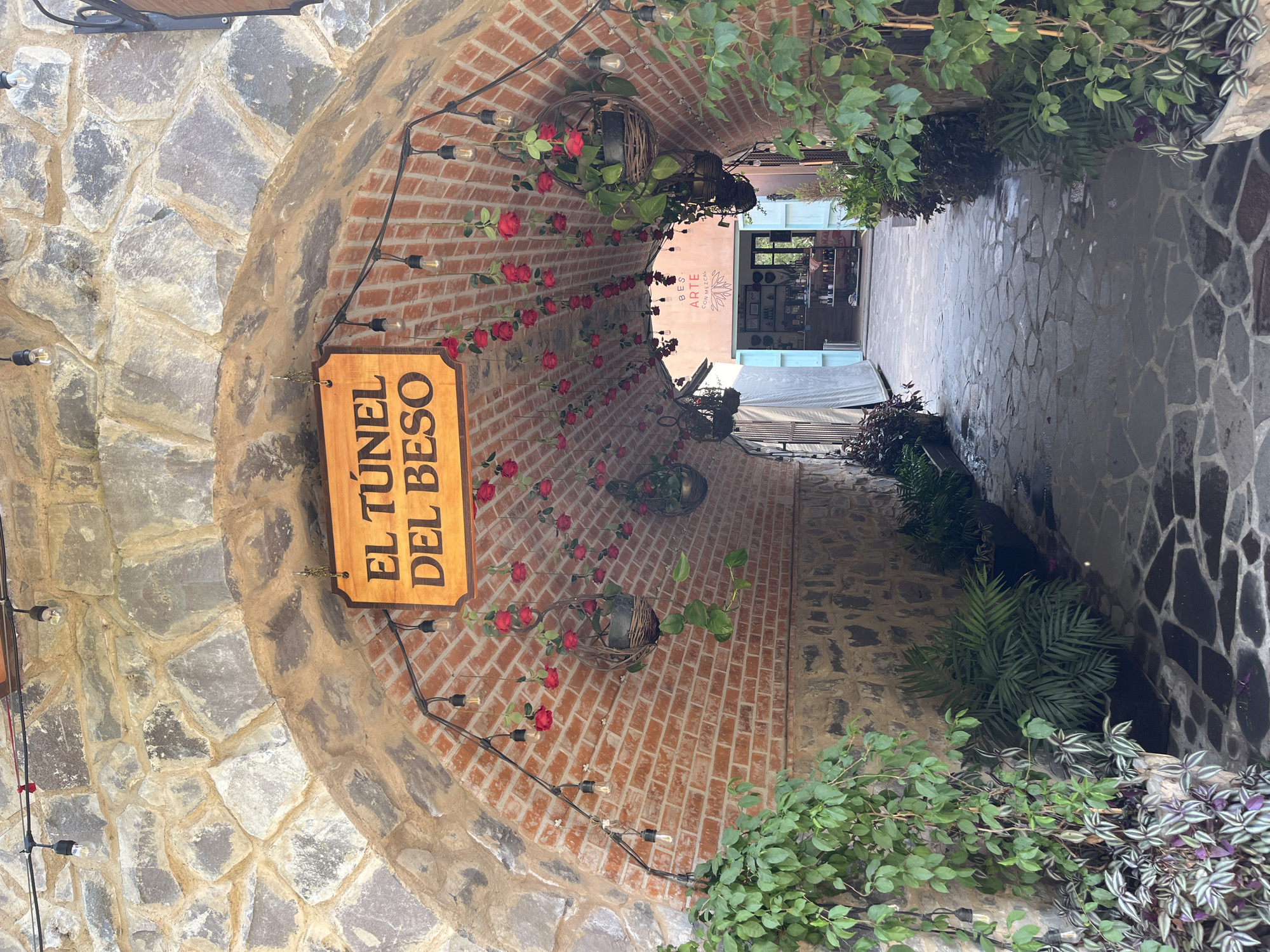 The image depicts a stone tunnel entrance, presumably in Domingo Arenas, Mexico, leading into a narrow passageway. A wooden sign reading "El Túnel Del Beso" is prominently displayed on the left side of the entrance. The tunnel's archway is constructed of stone and brick, with decorative red roses and hanging planters adorning the brick walls. String lights are also present along the arch. The passageway beyond the tunnel entrance is paved with irregular stone slabs, and plants grow along the walls. In the distance, a storefront with the word "ARTE" partially visible is seen at the end of the passageway. There are no people visible in the image.