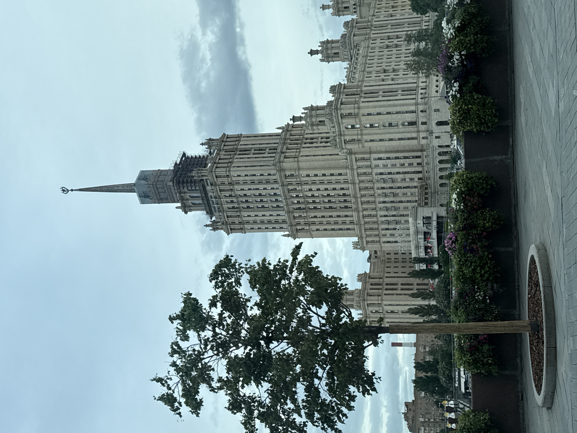 The image depicts the Kotelnicheskaya Embankment Building, one of the Seven Sisters skyscrapers in Moscow, Russia. The building is a prominent architectural landmark, characterized by its Stalinist architecture and towering spire. It is captured from a low-angle perspective, showcasing its height and grandeur. A tree with dense foliage occupies the foreground, partially obscuring the base of the skyscraper. The sky is overcast with cloudy conditions. A planter box runs along the right side of the frame, containing flowers. The area appears to be a public space with paved surfaces. There are no people visible in the photograph.