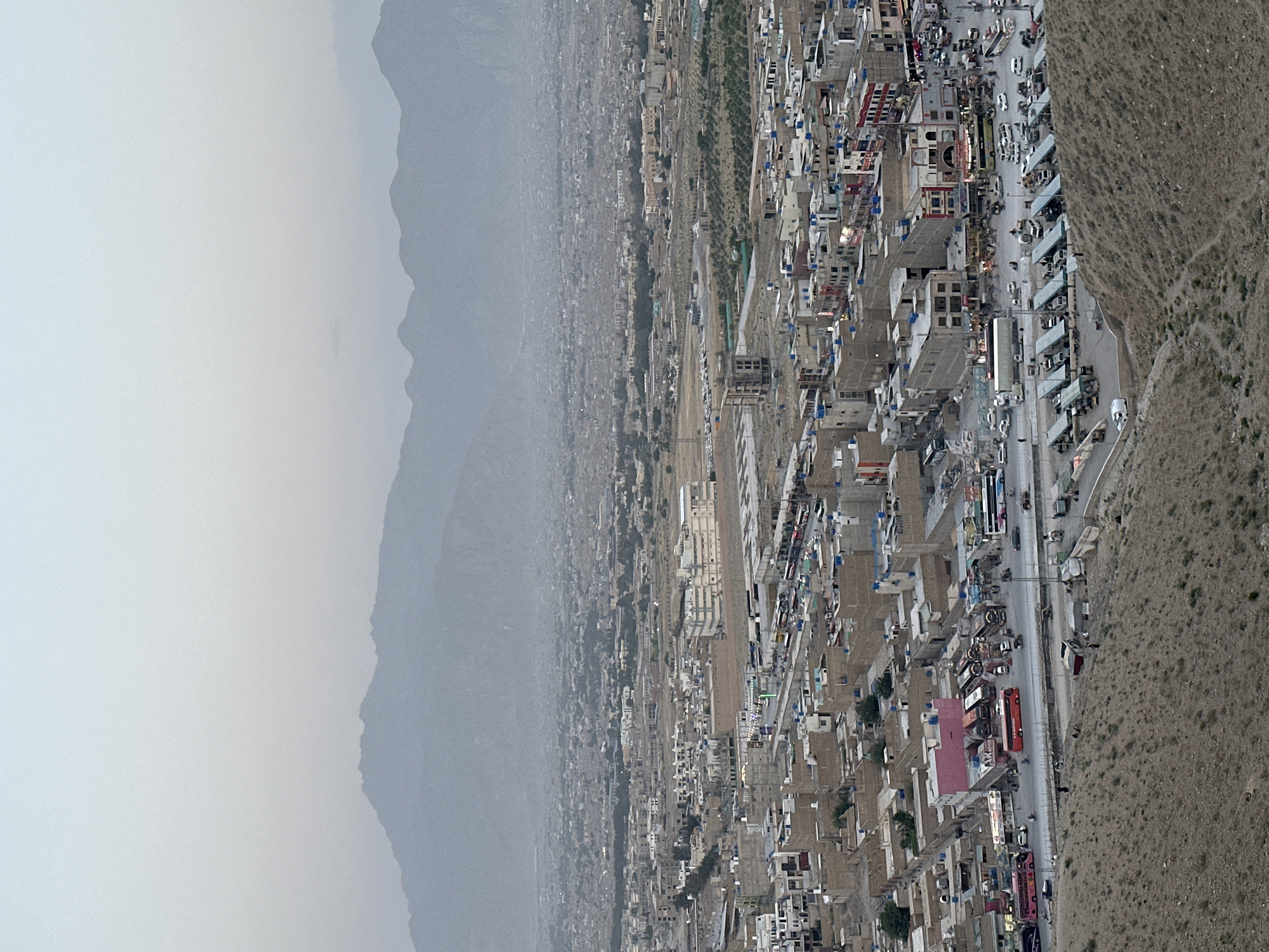 An aerial view shows a densely populated area along a highway in Quetta City Tehsil, Pakistan, adjacent to a mountain range. The architecture is a mix of low-rise buildings, with a significant number being uniform in style and color, suggesting a planned or standardized construction. A major road runs through the area with a line of commercial vehicles, likely buses, parked along the side, indicating a transportation hub. There is vehicular and pedestrian traffic on the road. The landscape transitions from the built environment to arid terrain on one side and mountainous terrain shrouded in haze on the other, highlighting the city's geographical context.