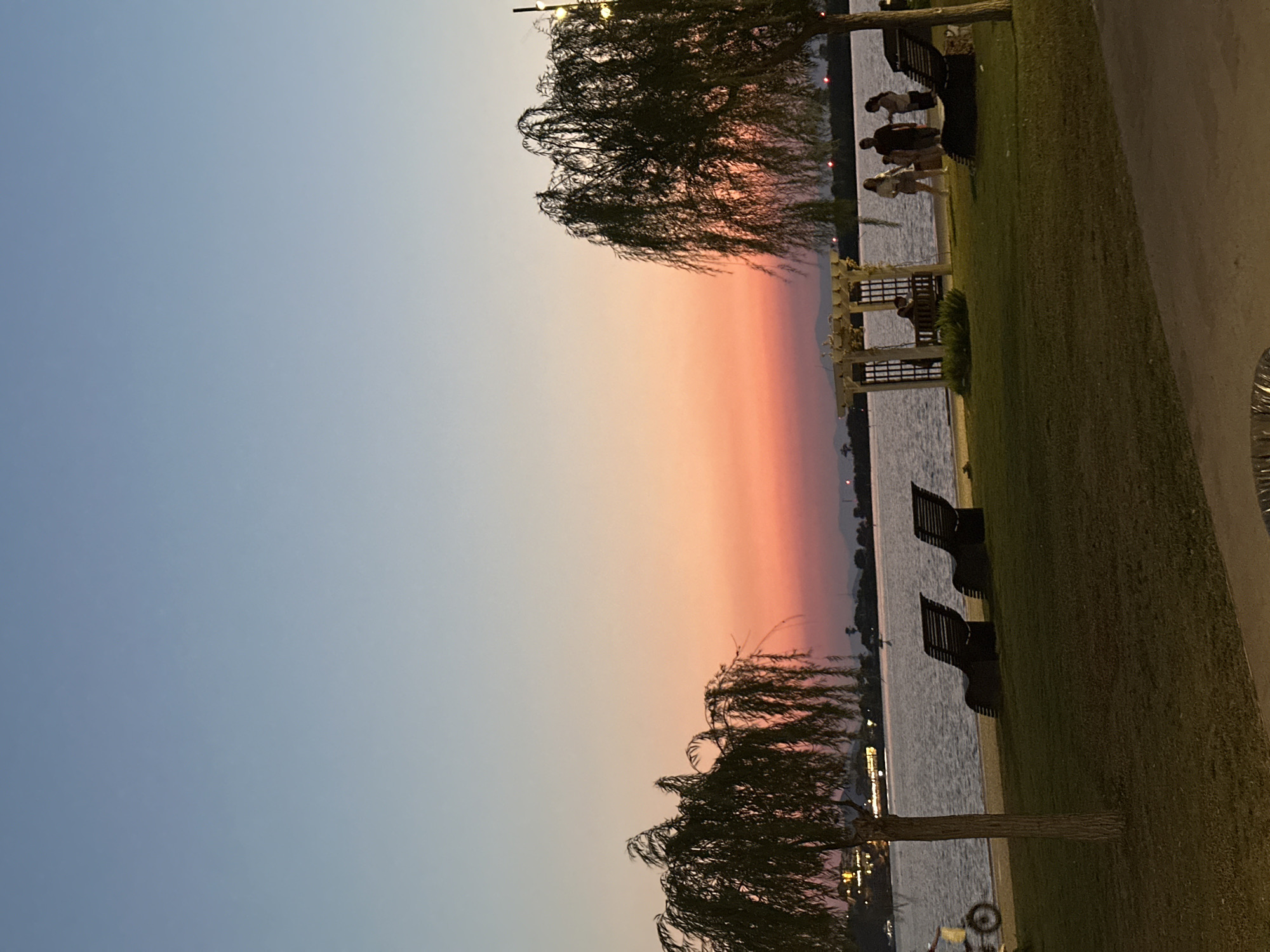 The image depicts a serene sunset scene at a waterfront location, likely in Fethiye, Turkey. The sky transitions from a pale blue at the top to a warm gradient of pink and orange near the horizon. In the foreground, a lush green lawn is dotted with several dark, modern lounge chairs. Two large weeping willow trees with their characteristic drooping branches frame the left and right sides of the scene.
The water, appearing calm, stretches across the middle ground, with a faint shoreline visible in the distance, possibly featuring some hills or mountains. On the lawn, a group of people is gathered near a wooden pergola structure. Three individuals, two women and a man, stand close together, appearing to be in conversation. Another woman is seated inside the pergola, and a man is also visible within the structure. To the left of the scene, a person is seen riding a bicycle on a paved path near the water's edge.
The overall atmosphere is tranquil and inviting, suggesting a public park or promenade area where people can relax and enjoy the evening. The presence of lounge chairs and the pergola indicates amenities for visitors. The warm hues of the sunset contribute to a peaceful and picturesque setting.
