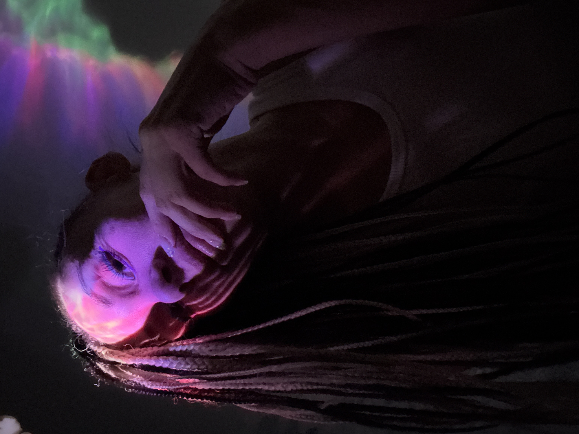 A person lies on their side with their face illuminated by pink and purple light. The person's right hand is held up to their face. Braids flow down from their head. A light display of green, purple, and pink is visible behind them. The location is unknown, and cannot be determined from the image.