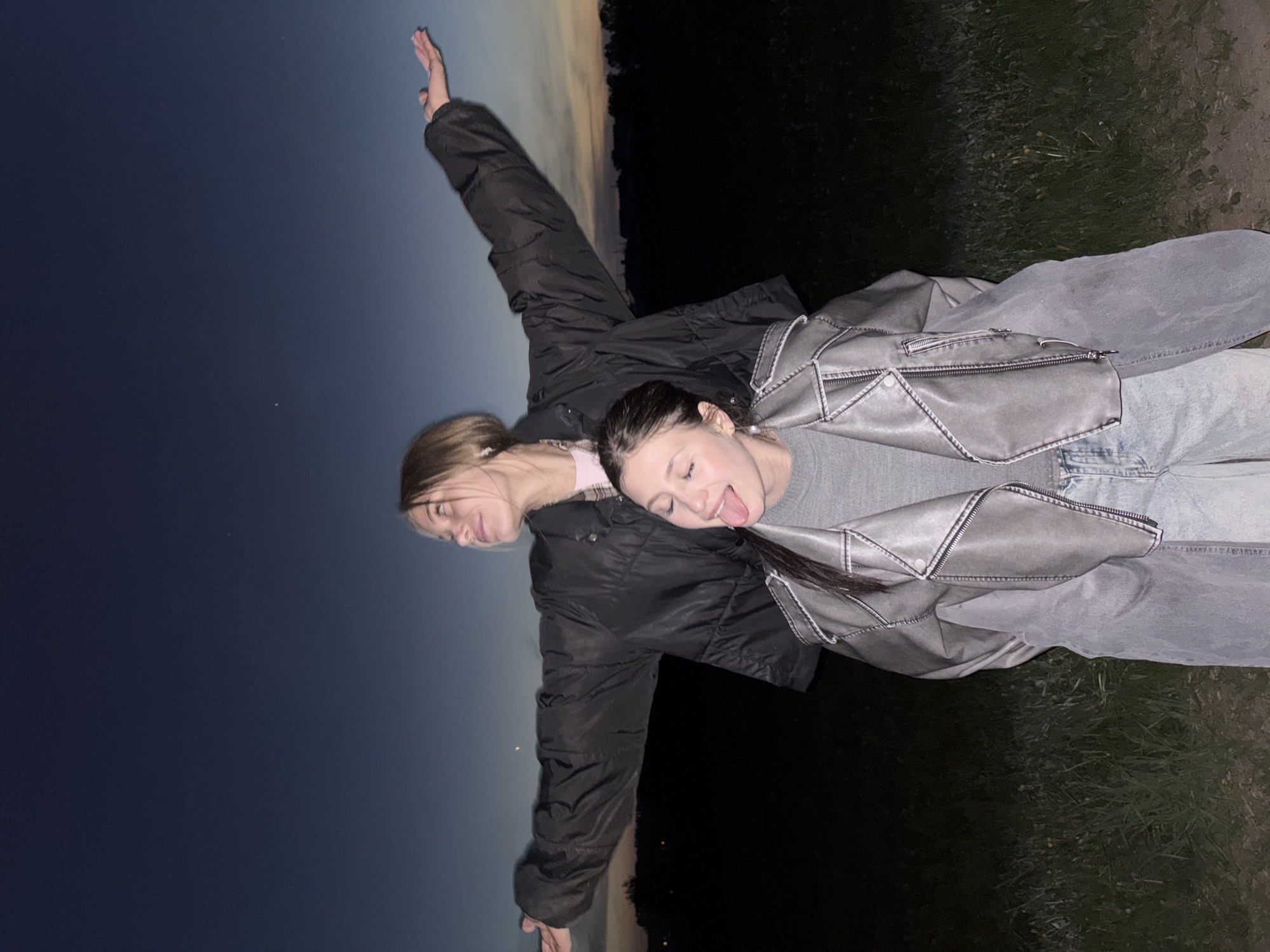 Two young women are posing for a photo outdoors. One woman is wearing a black puffy jacket, dark pants, and has her arm extended upwards. The other woman is wearing a gray leather-look jacket, gray shirt, and denim jeans. The photo is taken at dusk, with a dark blue sky in the background on one side and a dark, grassy area on the other. The location is not discernible to Фоки, Russia.