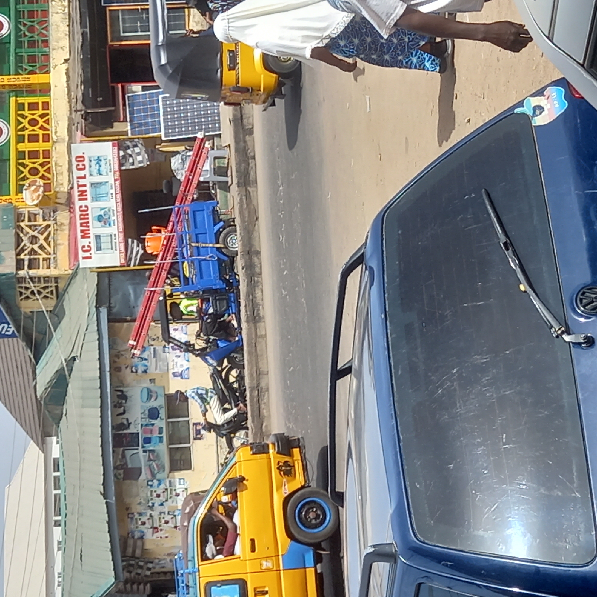 The image captures a vibrant, sunny street scene in what appears to be a commercial area in Sardauna, Nigeria. The view looks down a paved road lined with various vehicles and storefronts.
In the foreground, the rear of a blue car is visible on the right, featuring its back window, wiper, and a sticker that includes the text "RP-LS" and an illustration. The street itself is busy with several vehicles common to the region. A bright yellow auto-rickshaw (tuk-tuk) is present, with a person in a white garment and patterned blue attire walking alongside it, casting a distinct shadow on the pavement. Further along the road, a blue motorized tricycle, possibly a cargo vehicle, is carrying a long red ladder and an orange bucket. A yellow pickup truck with blue accents is also visible, with a person inside its cab.
On the left side of the street, various buildings house businesses. One structure features decorative green and yellow latticework. Another prominent storefront displays multiple items, notably several solar panels. A large sign for this business reads "I.C. MARC INT'L CO." with further text "MAIN OFFICE NO. 27 ALIYU DAN SARDAUNA ROAD KADUNA," providing a specific location cue.
The scene is brightly lit by clear daylight, indicating a midday or early afternoon setting, with harsh shadows adding to the sense of strong sunlight. The atmosphere is that of a bustling urban environment with ongoing commerce and daily activity.
