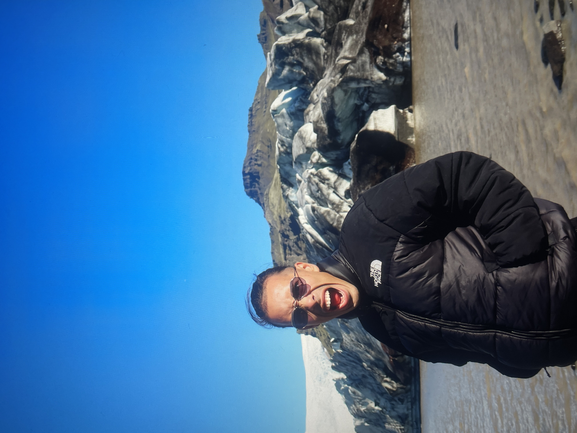 A person wearing a black puffer jacket and sunglasses is standing in front of a glacier. The person has their mouth open and appears to be yelling or shouting. The sky is clear and blue. The location is unknown.