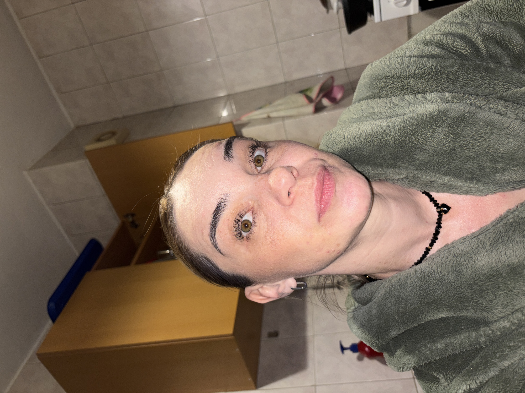 An adult female is depicted in a self-portrait taken indoors, specifically identified as a location within the city of Sinyavino, Russia. She has fair skin with visible freckles across her nose and cheeks. Her dark brown hair is pulled back, with a few lighter wisps visible near her forehead. Her brown eyes are wide open, gazing directly into the camera. She has distinct dark eyebrows and long eyelashes. Her lips are pink and slightly parted.
She is wearing a plush, fuzzy greenish-grey (sage green) bathrobe, covering her upper body. Around her neck is a black beaded choker-style necklace with a small, black, heart-shaped pendant. Silver-colored, rectangular-link hoop earrings are visible in both ears.
The background suggests a kitchen or utility room. The walls are covered in light-colored square tiles with visible grout lines. Two wooden, medium-brown overhead cabinets are present; one fully visible on the left and another partially visible on the right. A blue rectangular object rests atop the left cabinet. On the tiled wall to the right, a light-colored circular object (possibly a vent or light fixture) is present near the ceiling, and a patterned towel (primarily white with red/pink and green accents) hangs from a hook below it. In the lower left corner, a red pump bottle is partially visible. In the lower right, a black cooking pan rests on a white stovetop, part of a kitchen appliance, with a white control dial partially visible. The lighting is artificial and relatively dim, casting soft shadows.
