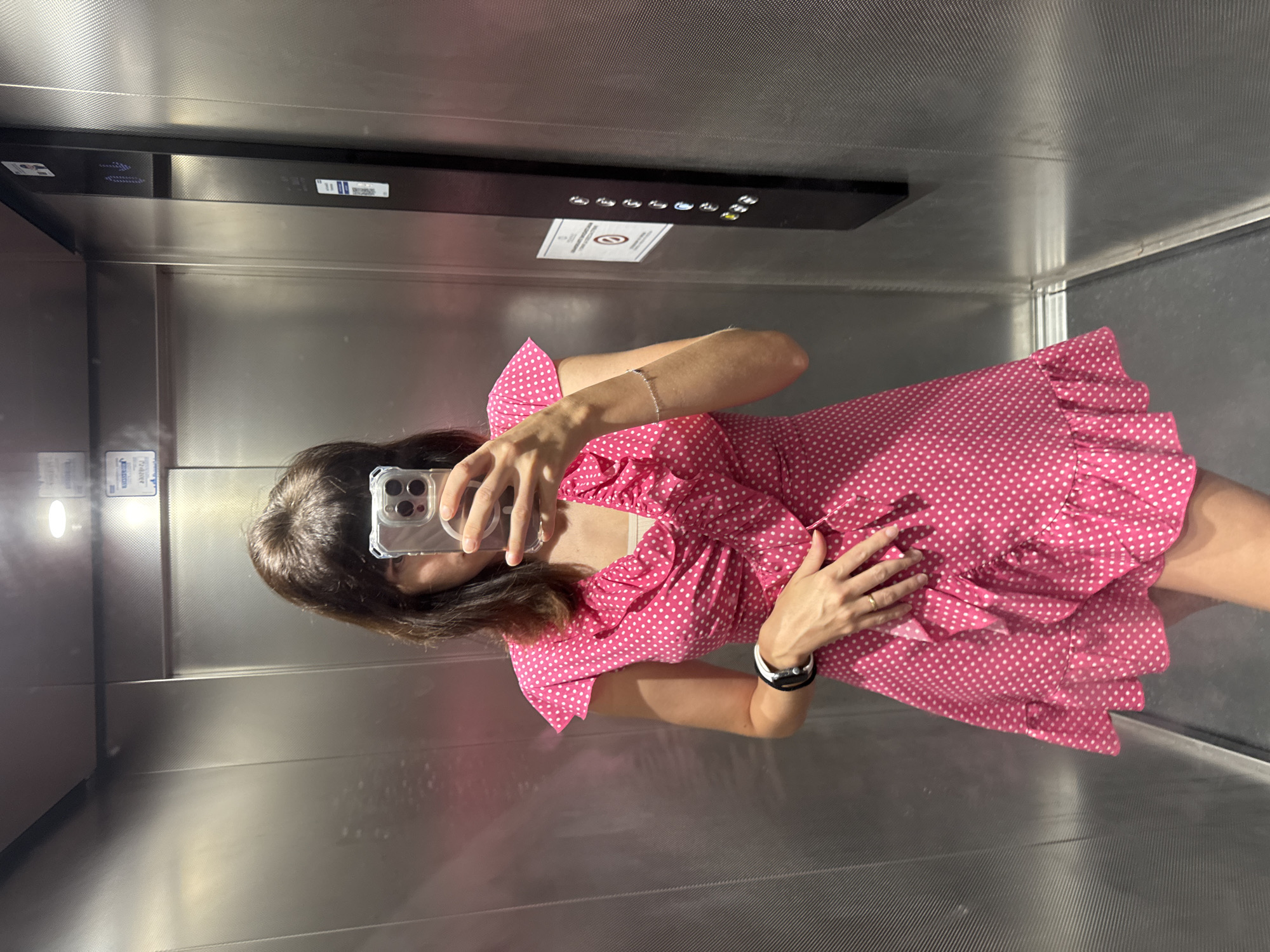 A young woman with shoulder-length brown hair, bangs, and fair skin is taking a selfie in an elevator. She is wearing a pink polka dot dress with ruffled sleeves and a V-neckline. Her left hand is placed on her stomach, and she is wearing a ring on her left ring finger. She has on a black and white bracelet on her left wrist and a delicate silver bracelet on her right wrist. The elevator walls are made of brushed metal, and the floor appears to be gray. On the right wall of the elevator, there is a black panel with illuminated buttons for the elevator controls, and a sign with a circular red symbol. The overall lighting in the elevator is bright and artificial, likely from the overhead lights. The scene appears to be captured in a modern, urban setting, possibly in Isola delle Femmine, Italy, given the elevator design and general aesthetic.