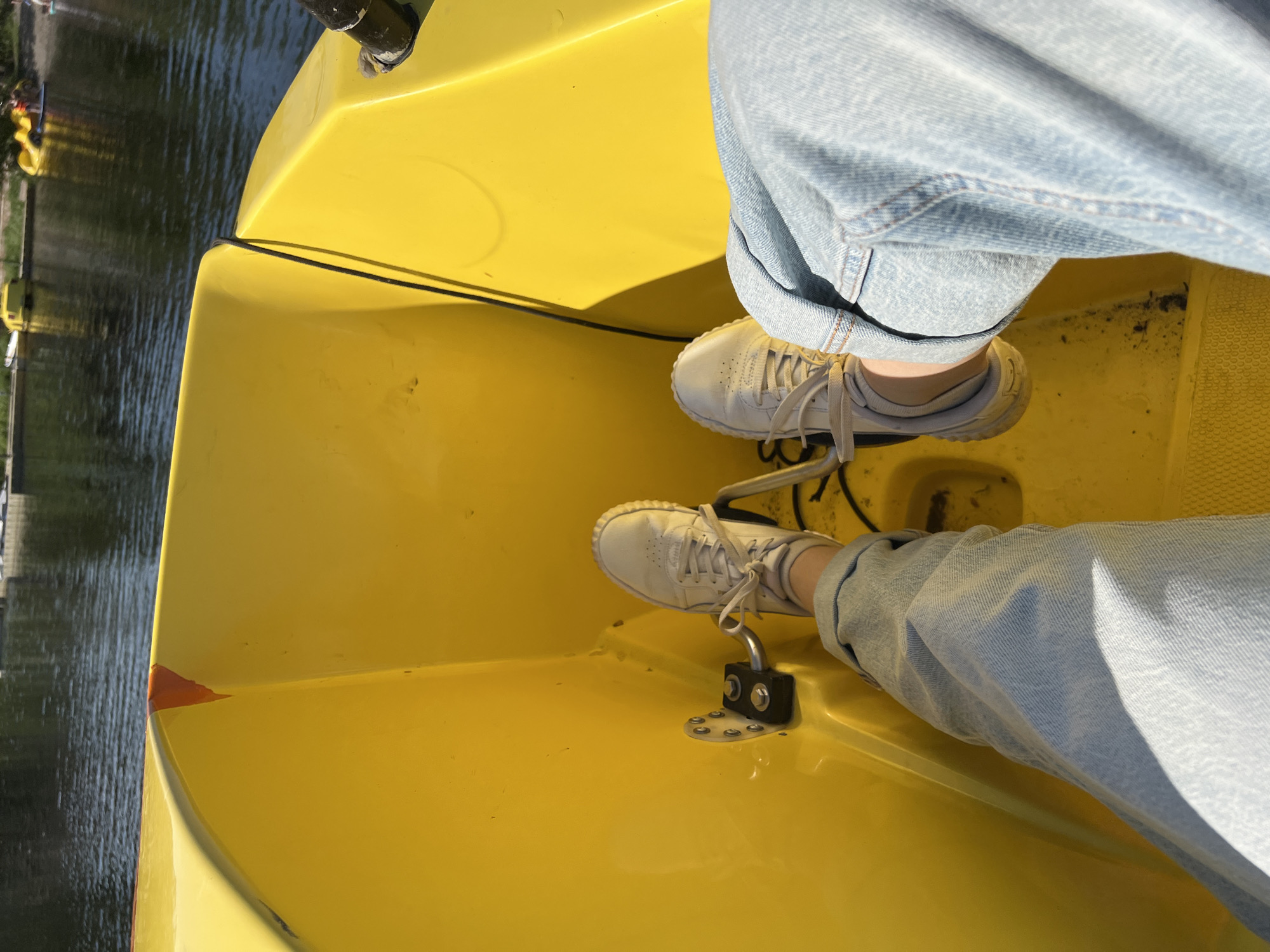 The image shows a first-person perspective from inside a yellow pedal boat on a body of water. The viewer's feet are visible, wearing light-colored sneakers and light-wash denim jeans. The pedals of the boat are positioned in front of the feet. The water in the background appears dark and rippling, with several other pedal boats visible near the shore. The shore is lined with trees and some buildings. The location is specified as Paratunka, Russia. The scene appears to be a sunny day.