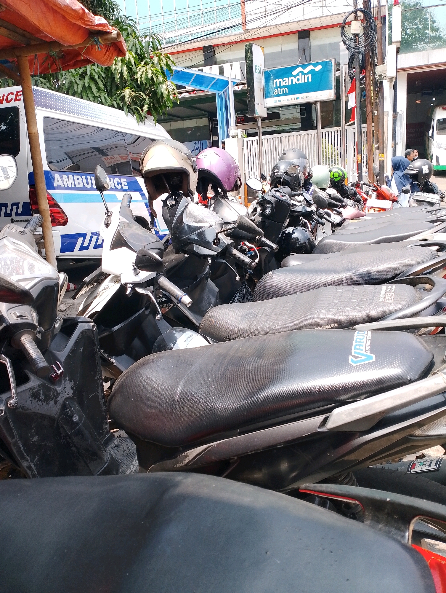 The image depicts a street scene in Bogor, Indonesia, likely in front of a building that houses a Mandiri ATM. In the foreground, several parked motorcycles are visible, with their seats and handlebars showing. One motorcycle in the center has a white and black body, and its side mirror is angled upwards. Another motorcycle to the left is silver and black. Helmets are placed on some of the motorcycles, including a gold and a pink helmet on the far left. Behind the motorcycles, an ambulance with "AMBULANCE" written in blue letters on its side is parked. The ambulance has a white body with blue and red detailing. To the right of the ambulance, a building with a blue sign that says "mandiri ATM" is partially visible, along with a metal fence. The environment suggests an urban setting with buildings and street infrastructure. The lighting indicates it is daytime.