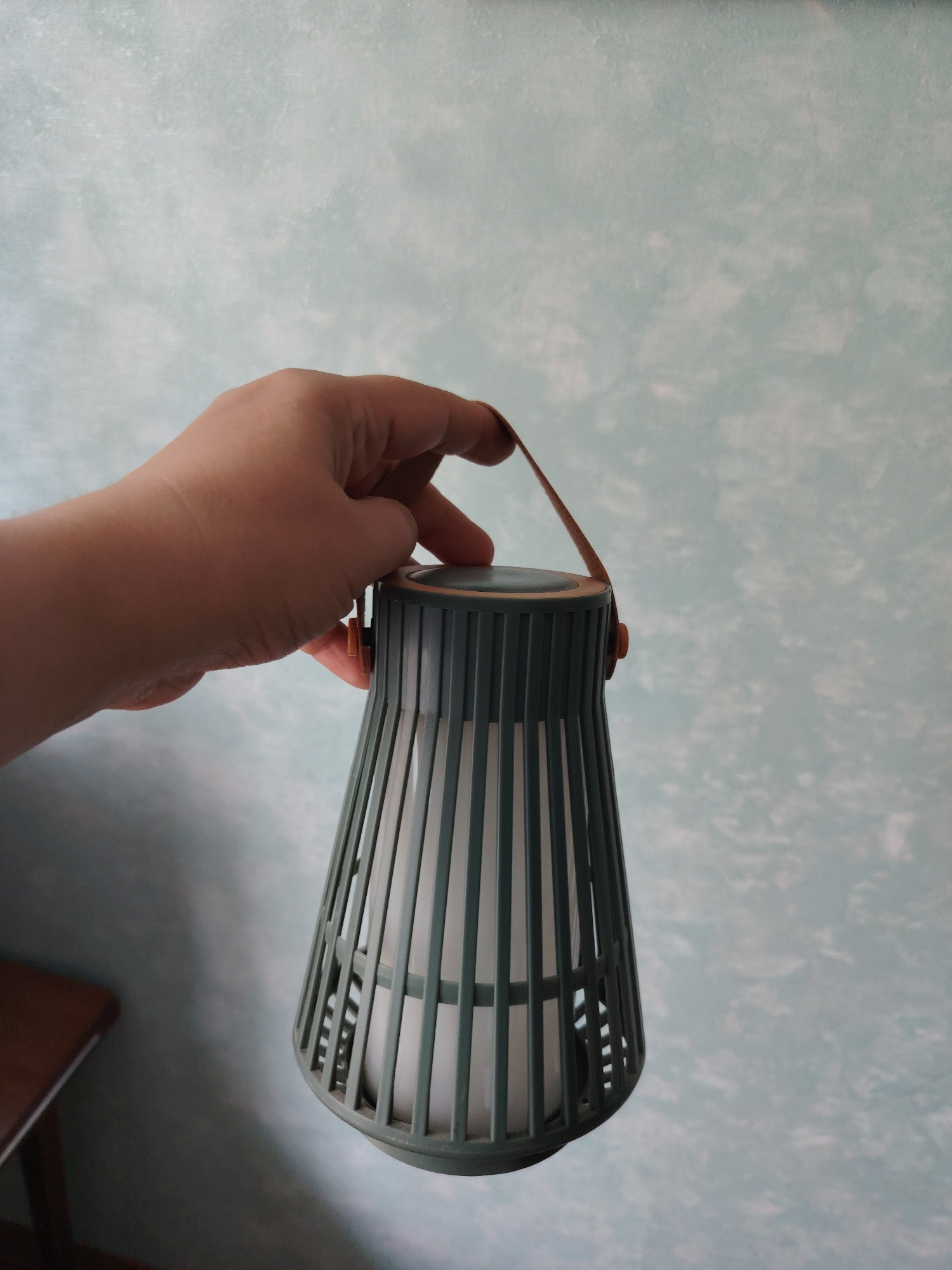 A hand holds a green lantern by its brown strap against a mottled blue and white wall. The lantern is cylindrical at the top and tapers towards the bottom. It has vertical slats and a white interior. A small part of a brown wooden table is visible in the lower left corner. The image is an indoor shot, and no specific location in the city of Volnoye, Russia can be determined from the visual information provided.