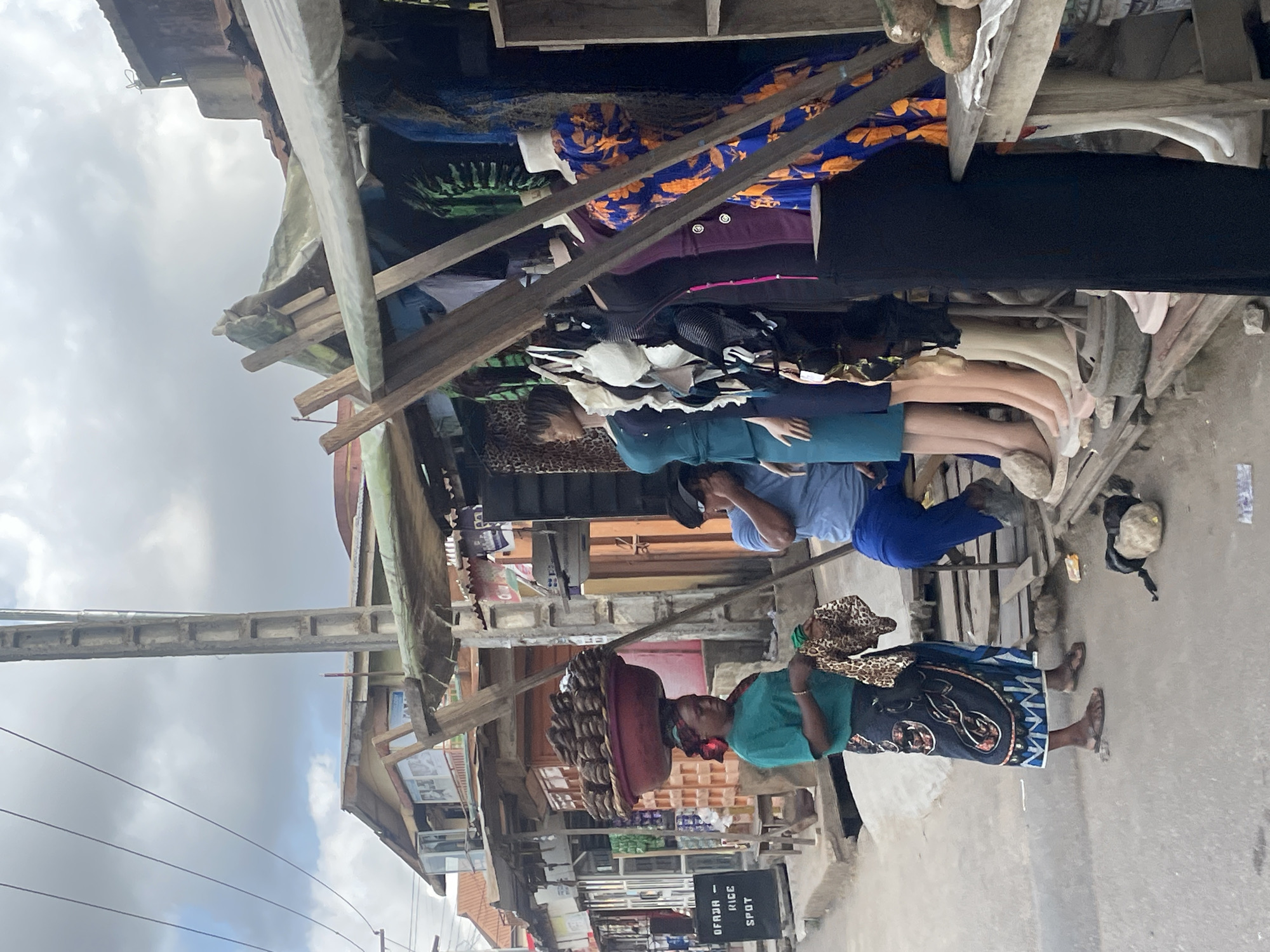 The image shows a street scene in Isheri, Nigeria. A woman is walking towards the right, carrying a large basket filled with food items on her head, and she is wearing a green shirt, a patterned skirt, and sandals. A second person, wearing a blue shirt and pants, is seen from the back and is facing towards the left. Market stalls line the street, showcasing various goods, including clothing and mannequins. The stalls are constructed with wood and covered with tarps, and some items hanging from the stalls. A sign indicating "OFAJA RICE SPOT" is visible. The street appears to be paved, and the sky is overcast. A concrete structure runs across the top left of the image.