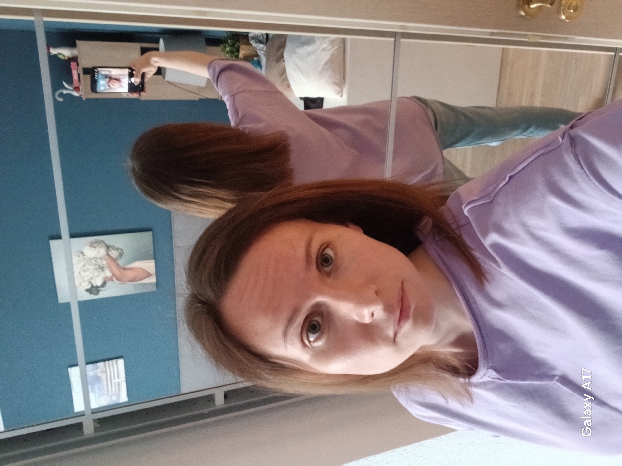 The image is a selfie taken in front of a mirrored closet. A woman is holding a smartphone to take the picture. Her face is prominent in the foreground, and her reflection can be seen in the mirror behind her. She is wearing a light purple t-shirt. The reflection shows her holding the phone with her right hand. In the background, visible through the mirror, is a blue wall with two pictures hanging on it. Other details visible in the reflection include a shelf with items on it, a hanger, and a pillow. The words "Galaxy A17" are visible in the bottom right corner. The location is not explicitly mentioned and there is nothing in the image that indicates it is Yekaterinburg, Russia.