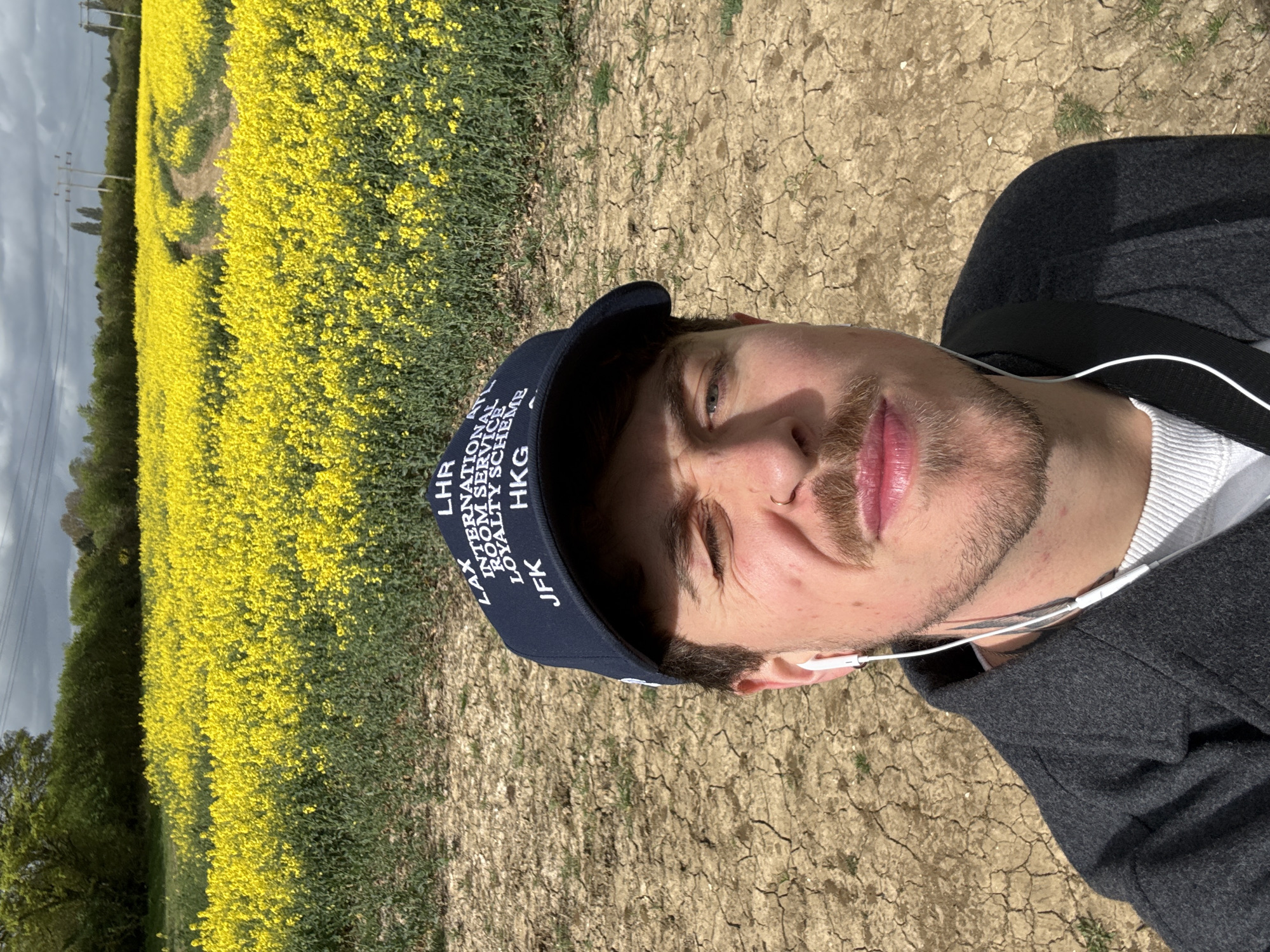 A man in a dark blue baseball cap with white lettering that reads "LAX LHR INTERNATIONAL ROOM SERVICE LOYALTY SCHEME JFK HKG" takes a selfie. He wears a gray jacket over a white shirt, and white earphones are visible. The background features a field of bright yellow rapeseed flowers, a dry, cracked earth path, and trees under a partly cloudy sky. While the man's location in North Hertfordshire, United Kingdom is mentioned, it is not visually evident within the image's content itself.