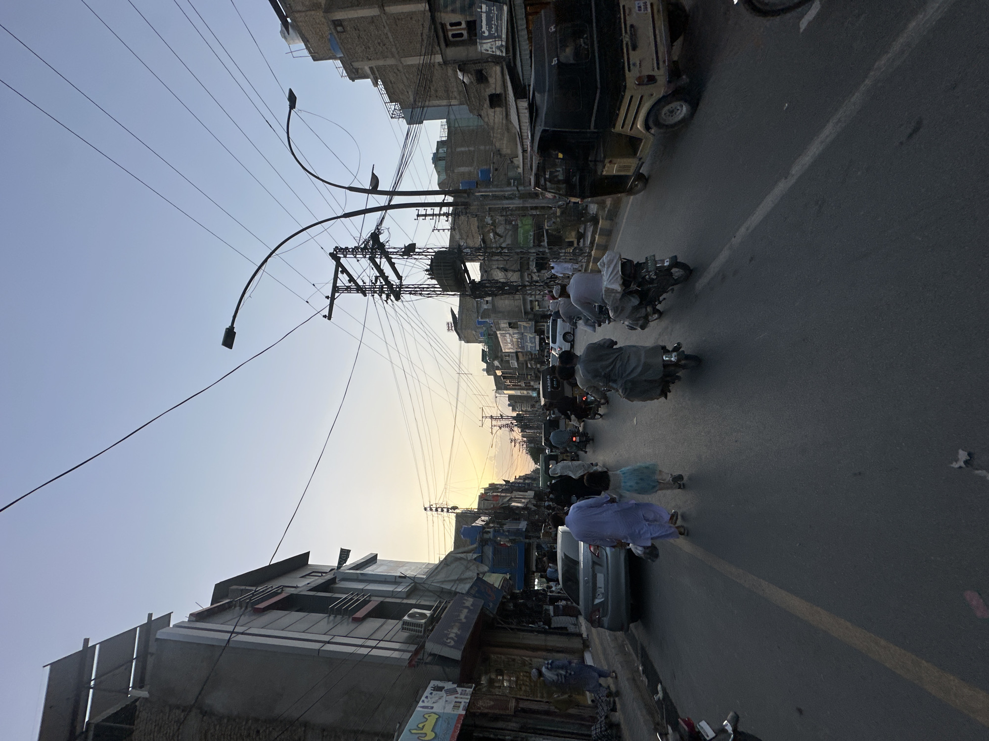 The image depicts a street scene in Quetta City Tehsil, Pakistan. Several individuals, some in traditional clothing, are walking or riding motorcycles on a paved road. Buildings line both sides of the street, with various shops and signs visible. Utility poles with a dense network of wires are prominent overhead. A vehicle, possibly a rickshaw or auto-rickshaw, is also present. The lighting suggests either sunrise or sunset, with a soft, warm glow on the horizon.