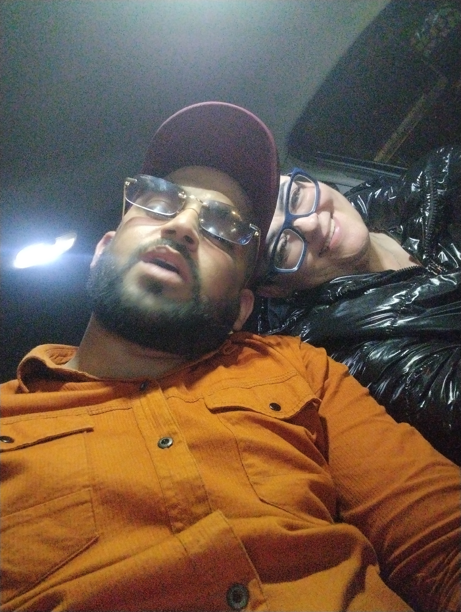 A selfie shows two individuals, one wearing an orange shirt and a maroon baseball cap, and the other wearing blue-framed glasses and a black, shiny puffer jacket. The subjects are likely inside a car, indicated by the interior lighting and the angle of the photograph. There are no immediate indicators in the photo that place it definitively in Martil, Morocco. Without additional context or visual clues, the location in Martil cannot be confirmed.