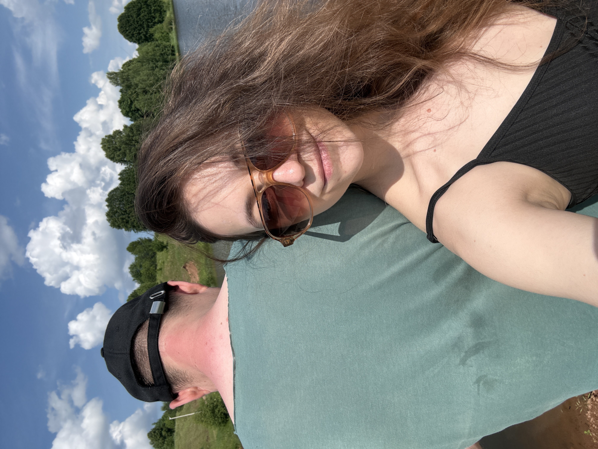 In Zasechnyi, Russia, a woman wearing sunglasses and a black tank top poses for a selfie in the foreground. Behind her, a man wearing a green t-shirt and a black baseball cap is visible from the back. The background features a body of water, a lush green landscape, and a bright blue sky with scattered clouds. The overall scene suggests an outdoor recreational activity, possibly near a lake or river.