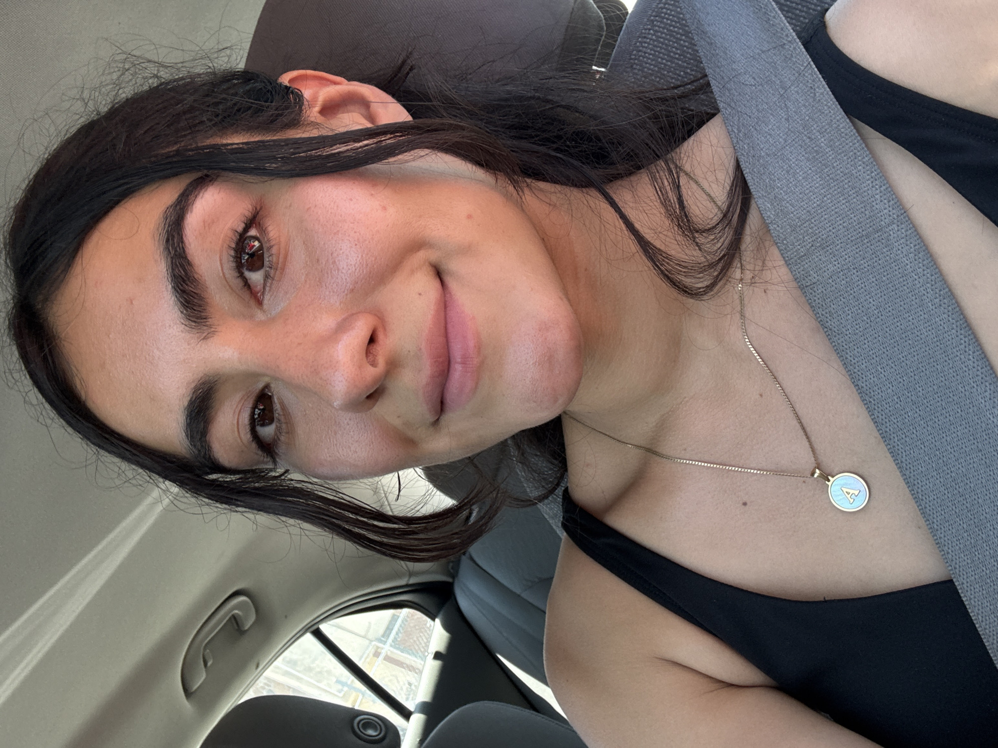 The image features a young woman in a car, captured in what appears to be a selfie taken during daylight hours in Puebla, Mexico. She has long, dark hair, dark eyes, and prominent, dark eyebrows. Her head is tilted, and she offers a subtle, closed-mouth smile, conveying a calm and content expression.
She is wearing a black tank top, and a gray seatbelt diagonally crosses her chest. Around her neck, she wears a delicate gold chain necklace with a notable round pendant. The pendant has a light blue, possibly mother-of-pearl, background with a distinct white capital letter "A" centered on it.
The setting is the interior of a car, with light-colored (possibly gray or beige) seats and ceiling visible. A car handle is discernible on the ceiling above her head. Bright light streams in from outside the car, visible through the windows, indicating it is daytime. The background outside the car window is blurred, showing indistinct shapes that could be buildings or structures.