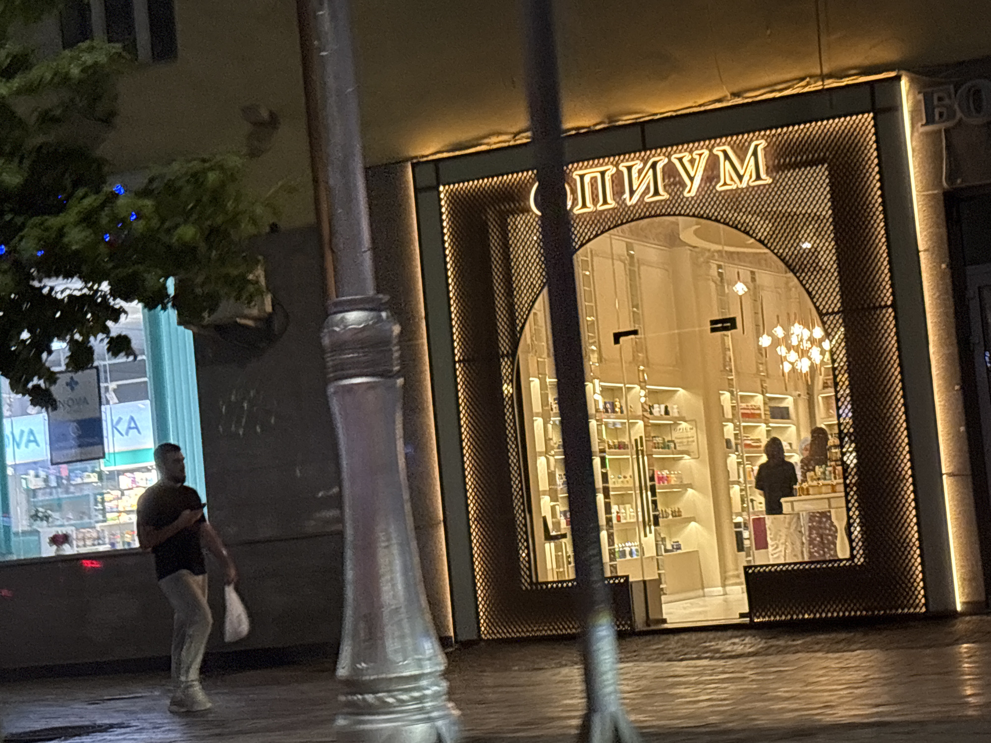 A street scene at night in Grozny, Russia, captures a storefront with illuminated signage that reads "ОПИУМ" in large, gold lettering. The storefront has a large arched window showcasing shelves stocked with various products, possibly cosmetics or perfumes. Two individuals, their figures indistinct due to shadow and distance, are visible inside the store. To the left of the store, a man walks on the sidewalk carrying a white plastic bag and looking at his phone. Another building with windows is visible behind him, with blue lights decorating a tree in front of it. A lamppost stands prominently in the foreground, partially obscuring the view of the store. The ground appears wet, reflecting the ambient light. The overall impression is of a modern retail environment on a city street after dark.