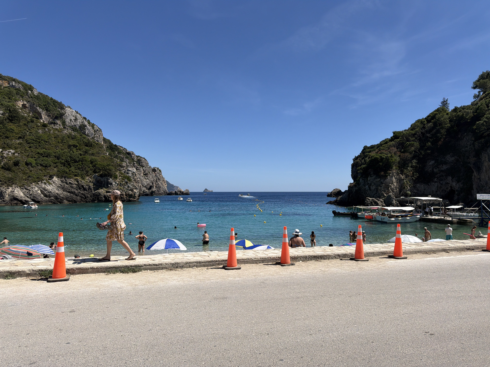 The image depicts a sunlit coastal scene in Palaiokastritsa, Greece, characterized by a blue sky, clear turquoise water, and verdant cliffs.
In the foreground, a light grey asphalt road occupies the bottom third of the frame. Six bright orange traffic cones, each resting on a black rubber base, are evenly spaced along the road's edge, positioned on a narrow, light-colored concrete strip. This strip abuts a low, light-colored stone wall, approximately 0.5 to 0.7 meters high, which separates the road from the beach area.
On the beach and in the water's edge, a woman wearing a patterned dress and a wide-brimmed hat walks from left to right along the stone wall, carrying sandals in her left hand. Two beach umbrellas are visible: one with blue and white stripes to the left, and another with blue and yellow stripes further right. Approximately 10-15 individuals are present in the shallow, clear water, swimming, wading, or standing. A pink inflatable tube is visible near one individual in the water. A yellow floating rope with buoys defines a swimming area further out.
The midground features the calm, blue-green water of a cove. Several white motorboats are anchored or moored within the cove. On the far right, a small dock extends into the water, accommodating multiple larger tour boats, some with white canopies, and approximately 5-10 people are visible on or near this dock.
The background is dominated by steep, rocky cliffs covered in dense green vegetation, framing the cove on both the left and right sides. These cliffs open to the wider, deeper blue sea beyond. A small, dark island or rock formation is discernible on the distant horizon in the center. The sky above is a clear, bright blue with minimal, wispy clouds in the upper right. A faint white contrail from an aircraft is visible in the upper left quadrant of the sky.