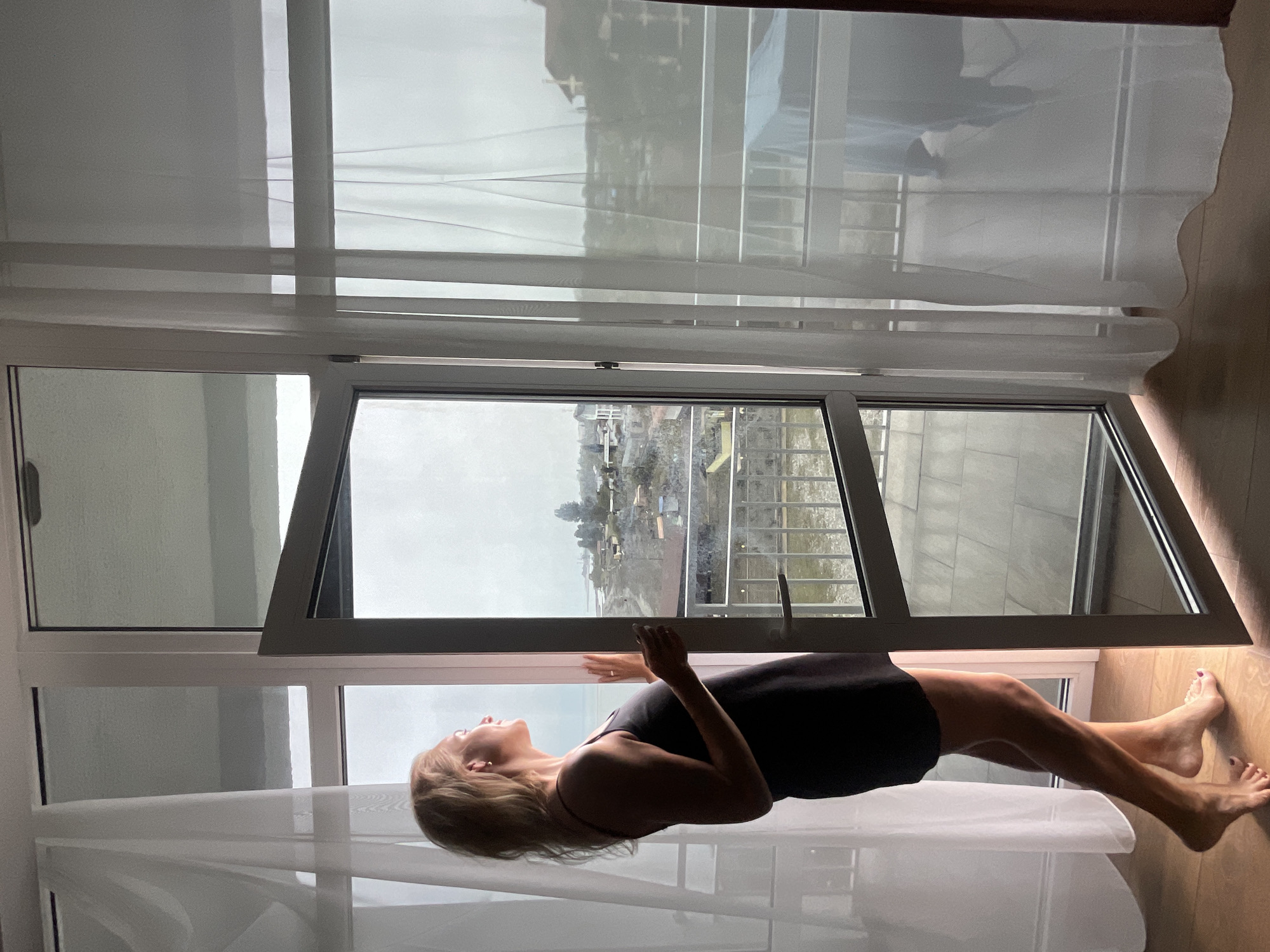 A medium, indoor shot presents a woman in a dark mini-dress. She is positioned lengthwise against an open window frame. The woman is leaning against the edge of the window; she is light-skinned and has short blonde hair. The woman has one arm extended, resting it on the frame.
The window reveals a grey sky, as well as buildings, trees, and a body of water. The environment outside the window is Giachrypsh, Abkhazia. The window is set within a white frame and is partially covered by sheer white curtains. The floor is wood and reflects the sunlight coming through the windows.