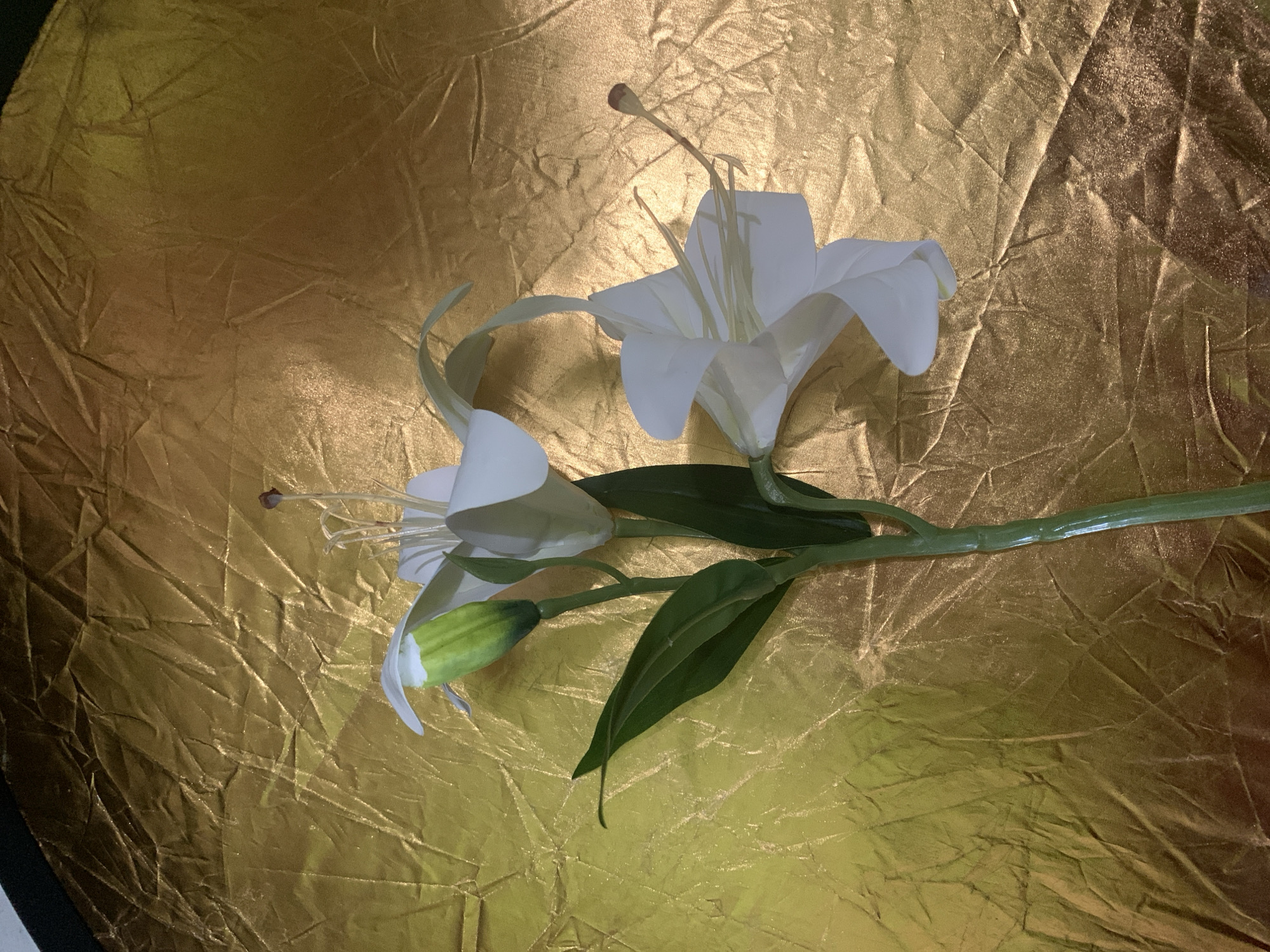 The image displays two white lily flowers and one lily bud with green leaves against a backdrop of crinkled gold fabric. The flowers are artificial, with visible seams and a slightly waxy appearance. One flower is fully open, revealing delicate white petals and yellow stamens. The other flower is partially open, and a single bud is closed. The stem is green and appears to be made of plastic. The gold fabric is illuminated, creating highlights and shadows that emphasize its textured surface. The lighting is somewhat harsh, causing reflections on the fabric and some overexposure on the flowers. There are no people present in the image, and no specific location in Kuala Pembuang, Indonesia, can be identified. The scene is a still life arrangement.