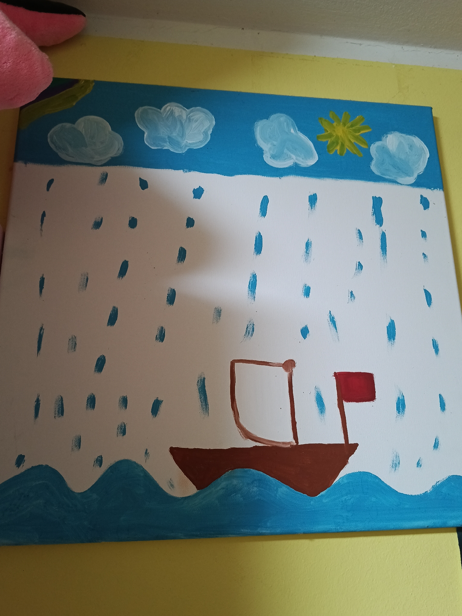 A child's painting is displayed on a yellow wall. The painting depicts a boat with a red flag sailing on blue waves under a white sky with blue raindrops. In the upper portion of the sky, there are white clouds and a yellow sun with radiating lines. A pink stuffed toy is partially visible in the upper left corner of the image. The painting appears to be on a canvas.