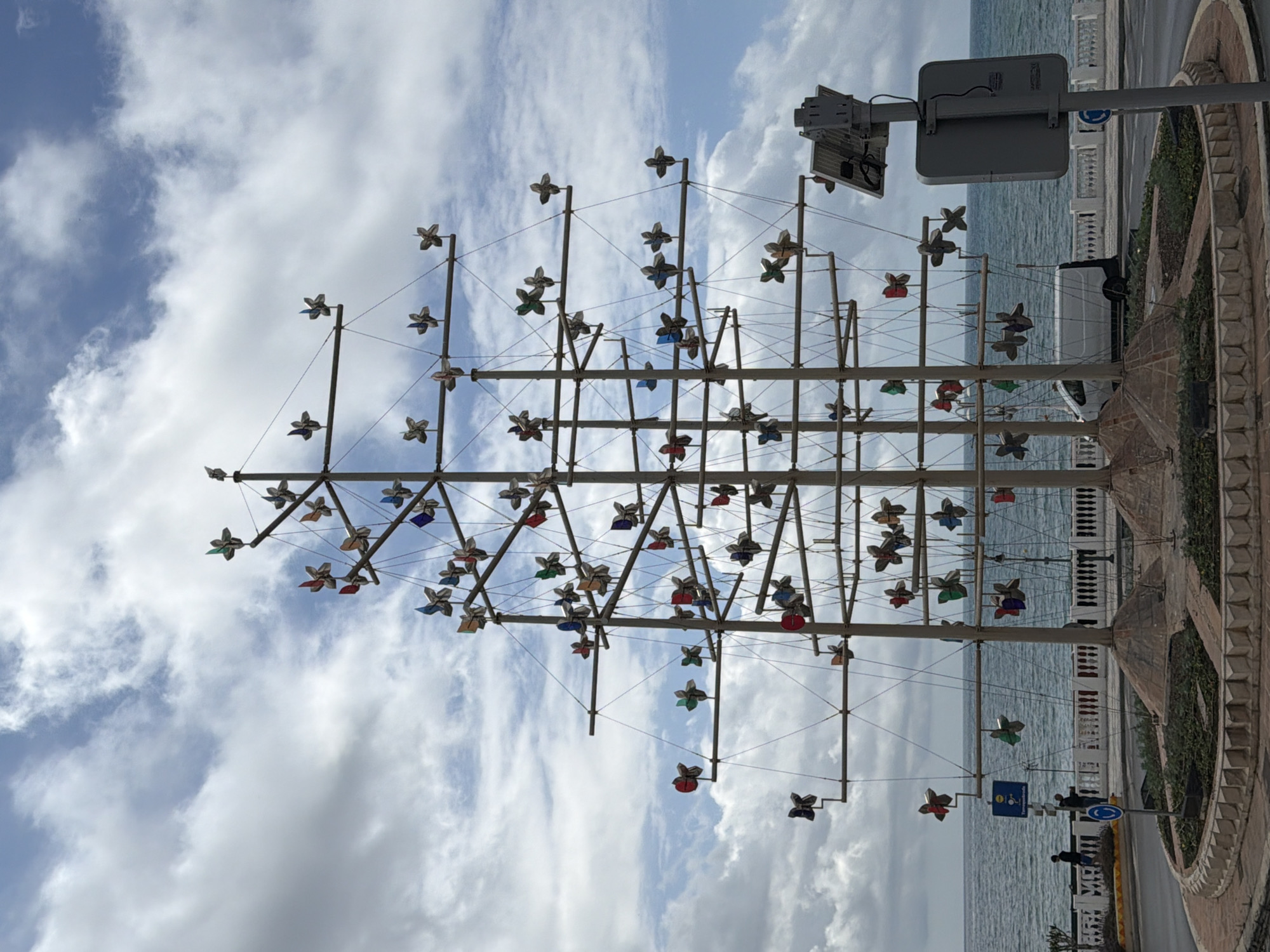 The image shows a metal sculpture resembling a ship's mast with numerous decorative elements resembling flowers hanging from it. The sculpture is positioned on a paved area, potentially a promenade, adjacent to the sea. In the background, the ocean extends to the horizon under a cloudy sky. Next to the sculpture are traffic signs and a street with a white van in the background. The location is likely Arroyo de la Miel-Benalmádena Costa, Spain, given the coastal setting and the style of the artwork and surrounding architecture. A person is walking along the street.