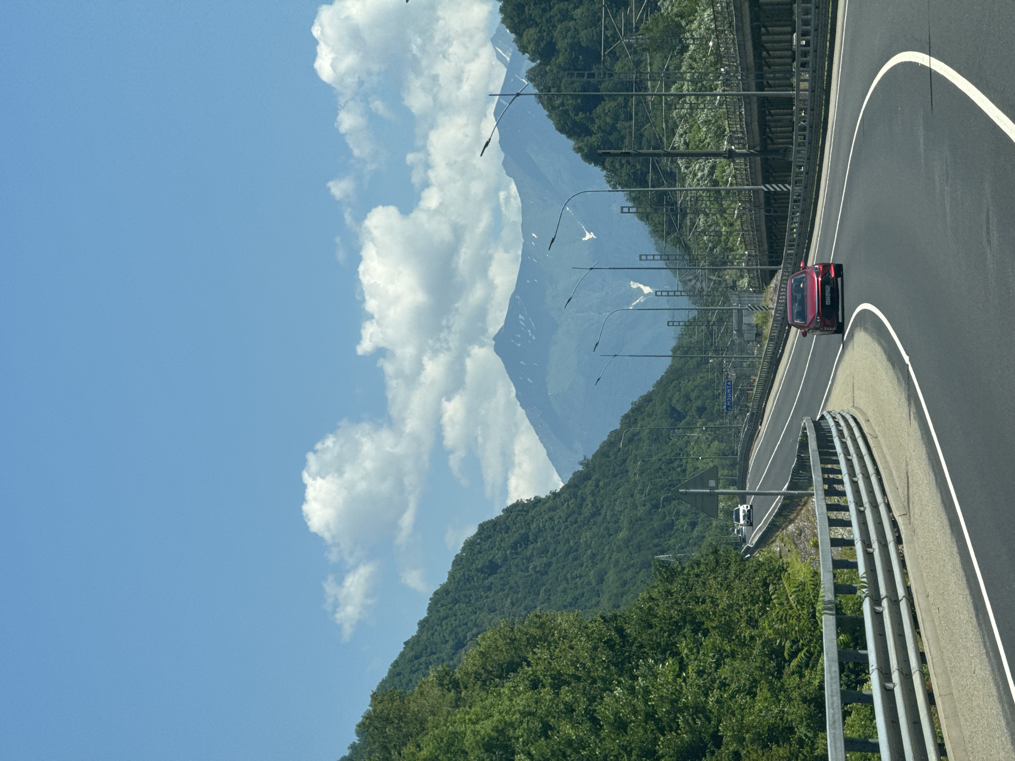 The image captures a view from a moving vehicle on a highway that curves through a mountainous, verdant landscape. The road, paved with asphalt, has clear white lane markings and a metal guardrail on the left. A red sedan is visible in the right lane, moving away from the viewer. The surrounding environment is characterized by lush green mountainsides densely covered with trees, and in the distance, snow-capped mountain peaks are visible under a bright blue sky dotted with white clouds. A railway line with overhead electrical wires runs parallel to the highway on the right. A sign with Cyrillic lettering, potentially indicating a location or direction, is visible in the background. The overall scene depicts a scenic drive through a mountainous region, likely in or around Chvizhepse, Russia, given the context.