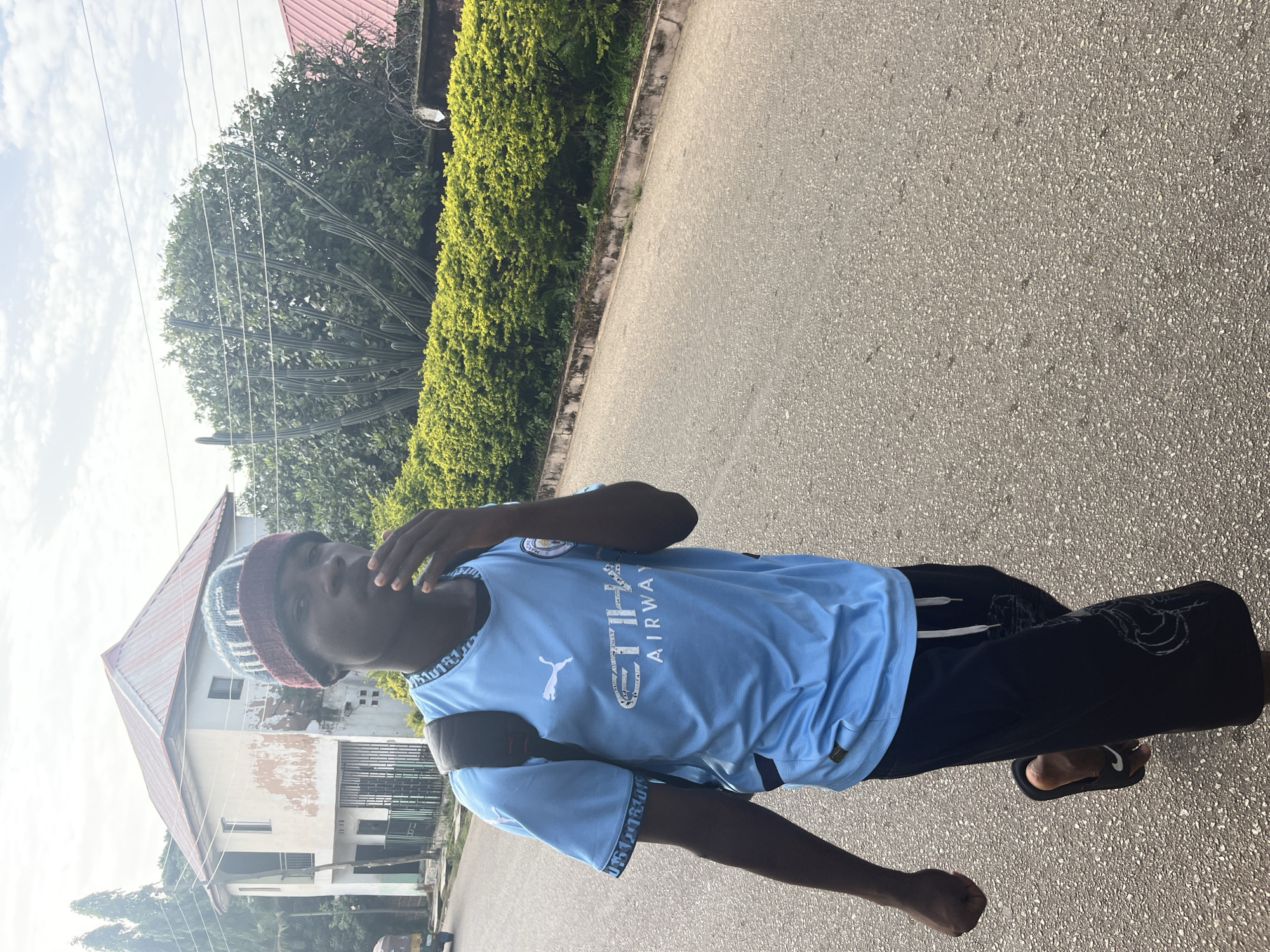 A young Black male walks on a paved road in Jos, Nigeria, carrying a black backpack. He wears a light blue Manchester City soccer jersey with a white Puma logo and "ETIHAD AIRWAYS" text, black shorts with white drawstrings and a white design, a multicolored knit cap, and black flip-flops. His left hand is near his mouth. In the background, a two-story building with a reddish-brown roof and peeling paint stands beside a dense green hedge and a tall, spiky cactus. The sky is overcast with scattered clouds.