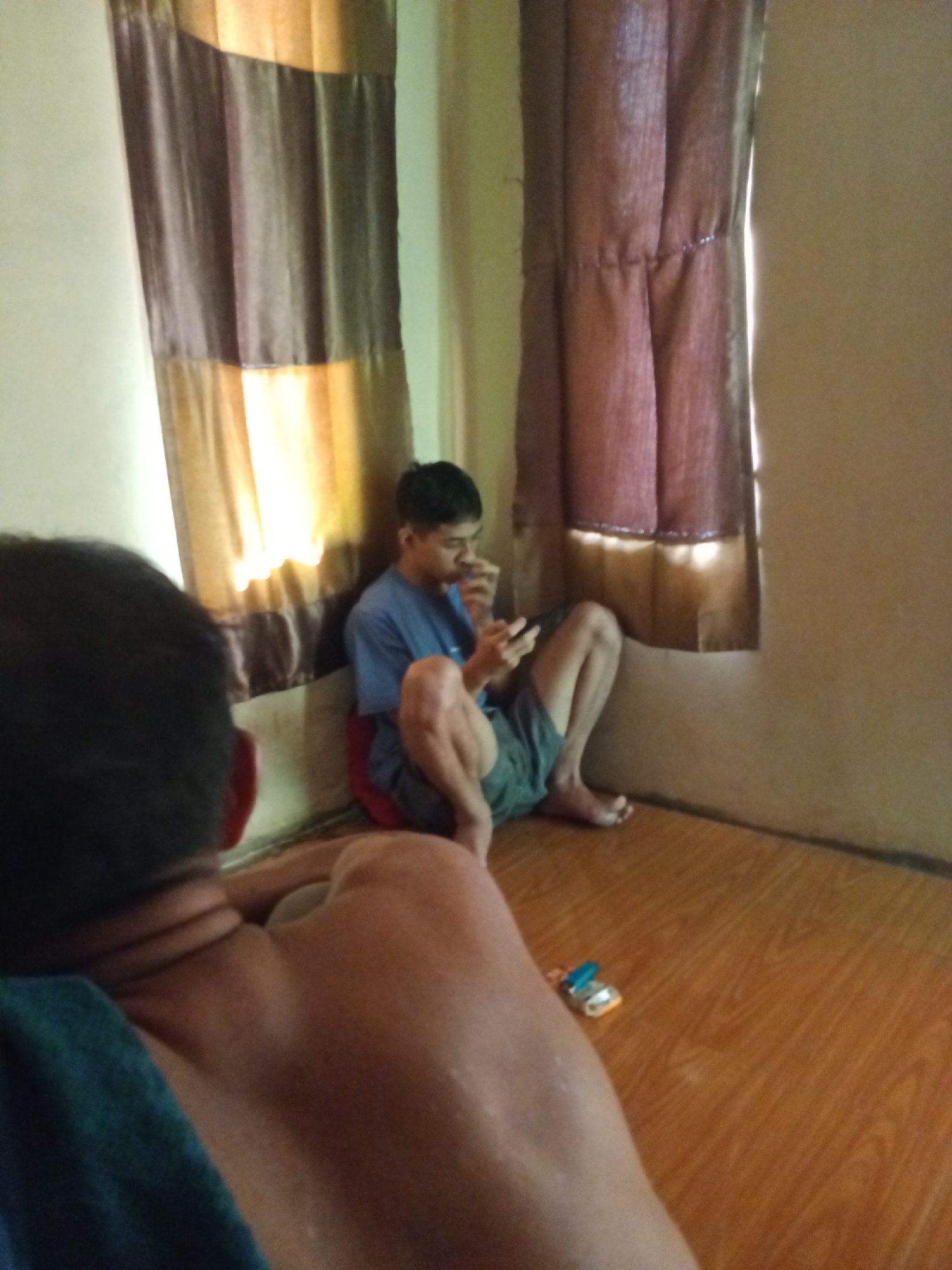 Two individuals are in a dimly lit room with wooden flooring and curtains. In the foreground, the back of a person's head and shoulders is visible. In the background, a young man in a blue t-shirt and gray shorts is seated on the floor, looking down at a mobile device. He appears to be eating or holding something to his mouth. The room has two windows covered by curtains, one a lighter brown and gold, the other a darker reddish-brown, both with reflective trim. A small toy is on the floor between the two individuals. The location is identified as Subang, Indonesia.