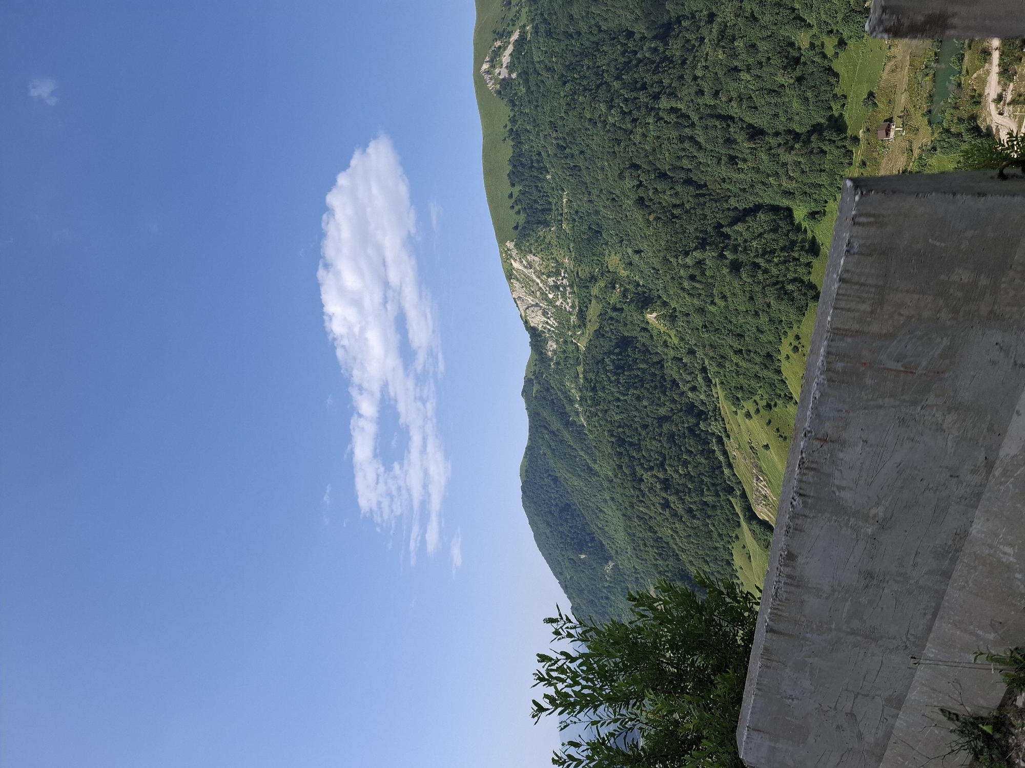 The image depicts a mountain landscape under a clear blue sky with scattered white clouds. In the foreground, a concrete barrier runs horizontally across the bottom of the frame. To the left, a cluster of green foliage obscures part of the barrier. Beyond the barrier, rolling hills covered in dense green trees dominate the midground. The trees form a thick canopy, with some patches of lighter green suggesting open fields or areas with less vegetation. Further in the background, a mountain peak with a rugged, rocky surface is visible. In the lower right portion of the image, nestled in a valley between the hills, there is a small red-roofed structure, possibly a house or cabin, situated near a body of water. The scene appears to be a natural, mountainous environment, likely in a rural area, with no people or animals visible.