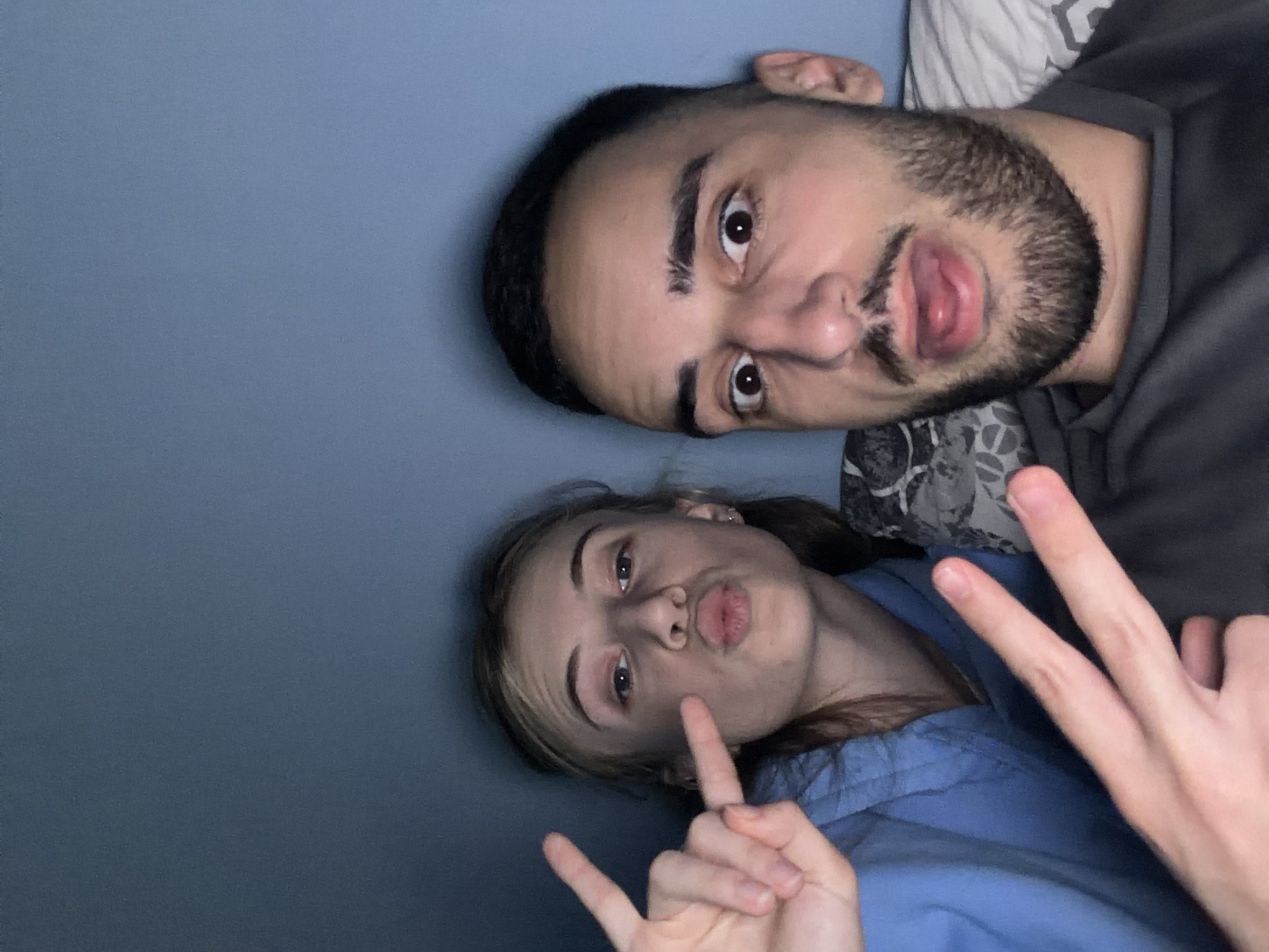 A selfie shot depicts two individuals against a dark blue background. The man is positioned at the top of the frame, with a dark beard and an expressive face. He is wearing a dark gray shirt, and a patterned pillow is visible behind him. The woman is below him, wearing a light blue top and making a "rock on" hand gesture with her left hand, while her right hand is making a peace sign. Her face is pursed in a kissing expression. The scene does not provide any visual information to suggest a specific location in Plovdiv, Bulgaria. The setting appears to be indoors, possibly a bedroom.