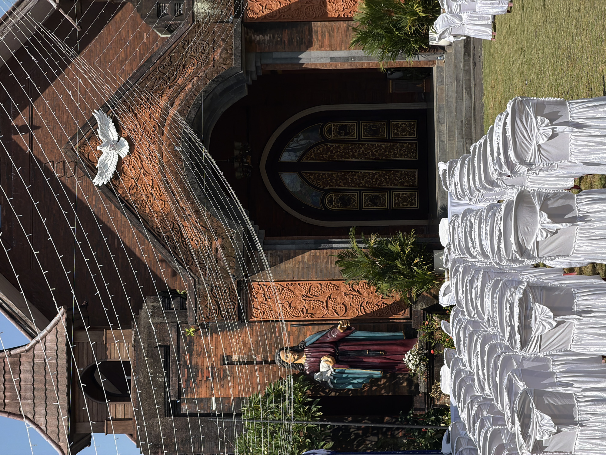In front of a brick church in North Kuta, Indonesia, rows of white-covered chairs are arranged on a grassy lawn. The church has a brown tiled roof and a large arched entrance with ornate doors. A statue of Jesus holding a lamb stands to the left of the entrance, and a white dove sculpture is mounted above it. String lights are draped across the facade of the building. The scene appears to be set up for an outdoor event.