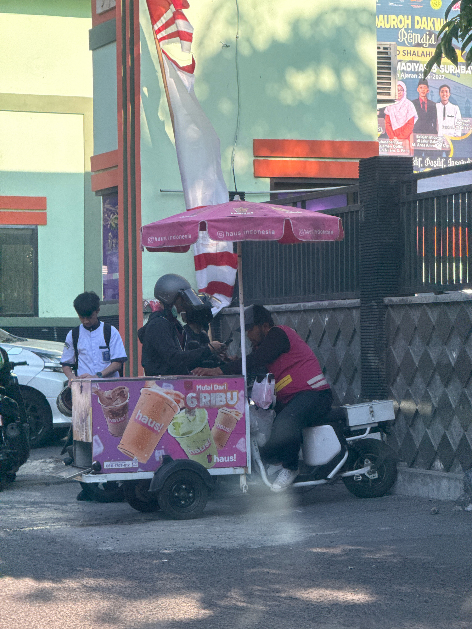 In Surabaya, Indonesia, a street vendor is operating a mobile drink stand. The stand is equipped with a pink umbrella featuring the "haus.indonesia" logo and displays images of beverages. Two individuals are present at the stand. One is a young man wearing a white uniform with a backpack, looking down. The other, who appears to be the vendor, is seated on a white electric scooter attached to the stand, wearing a pink vest and dark pants, and seems to be handling a phone. The background shows a building with a green and light blue facade, a prominent Indonesian flag, and a dark metal fence. The ground is paved, and sunlight casts shadows, suggesting an outdoor, daytime setting.